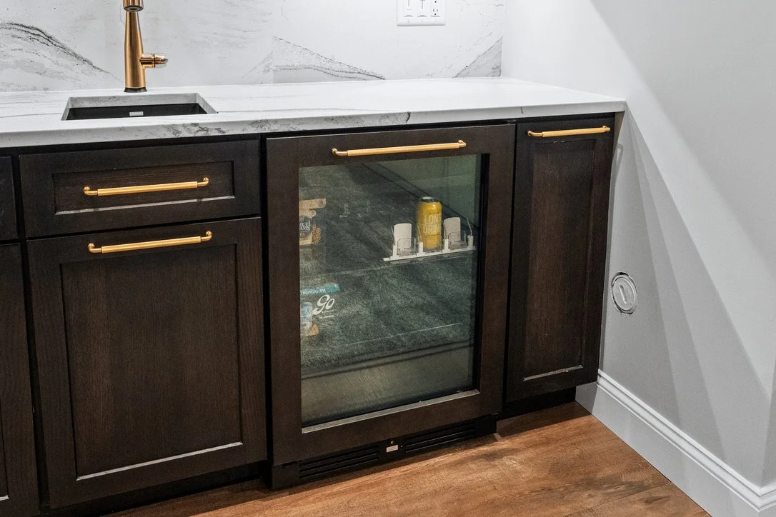 Basement bar with dark wood cabinets, marble backsplash, brass faucet, and a glass-front beverage fridge in a Brookville, Ohio basement renovation.