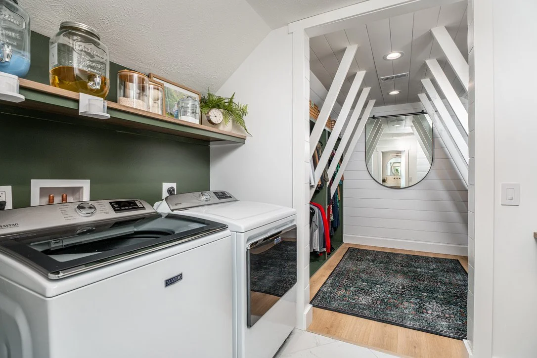 Laundry room with green accent wall, floating wood shelf, white top-load washer and dryer, and an adjoining shiplap closet with round mirror