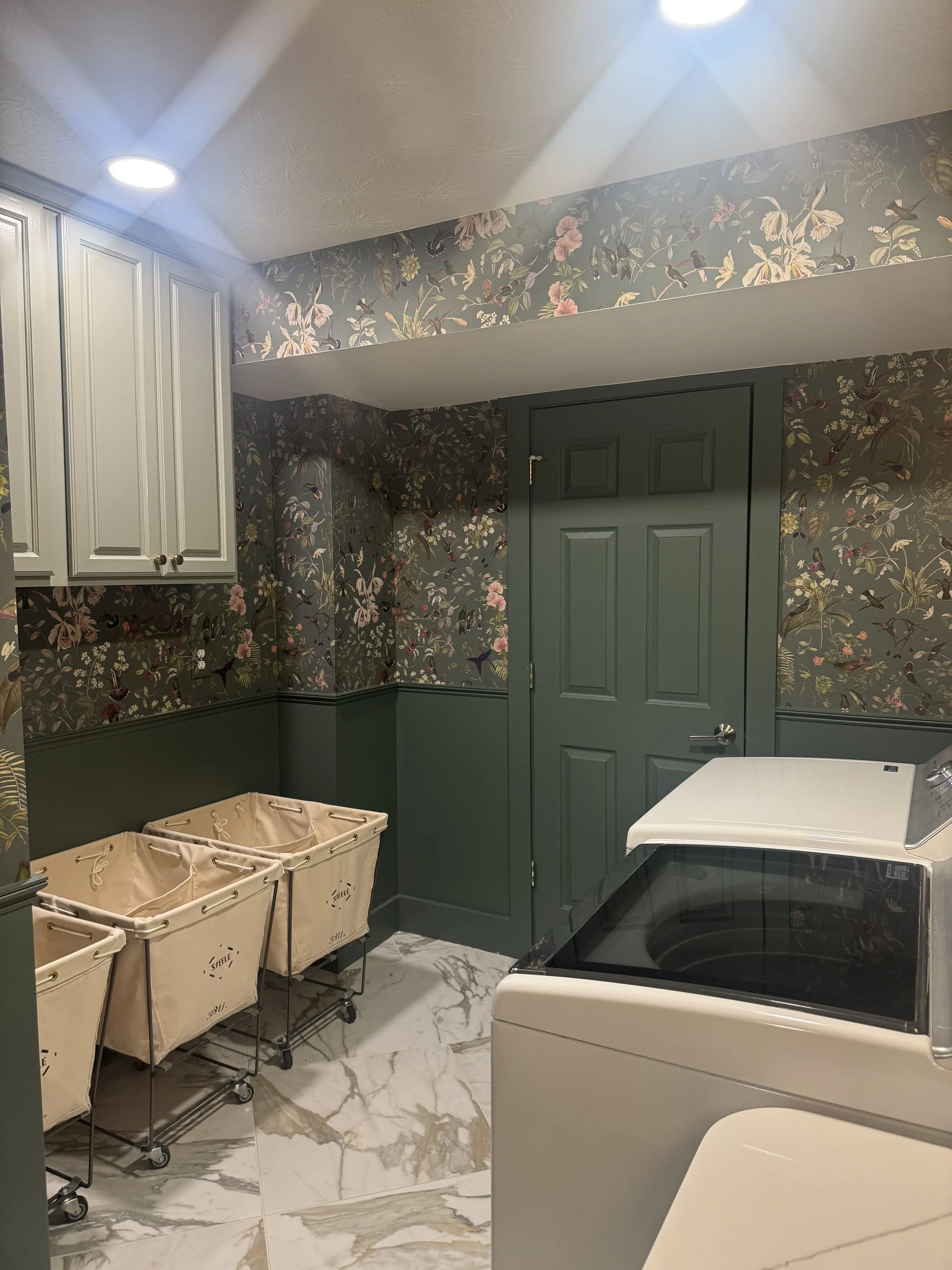 Beavercreek, Ohio basement laundry room renovation with canvas laundry sorters, upper cabinetry, green wainscoting, and botanical wallpaper by White Oak Interiors