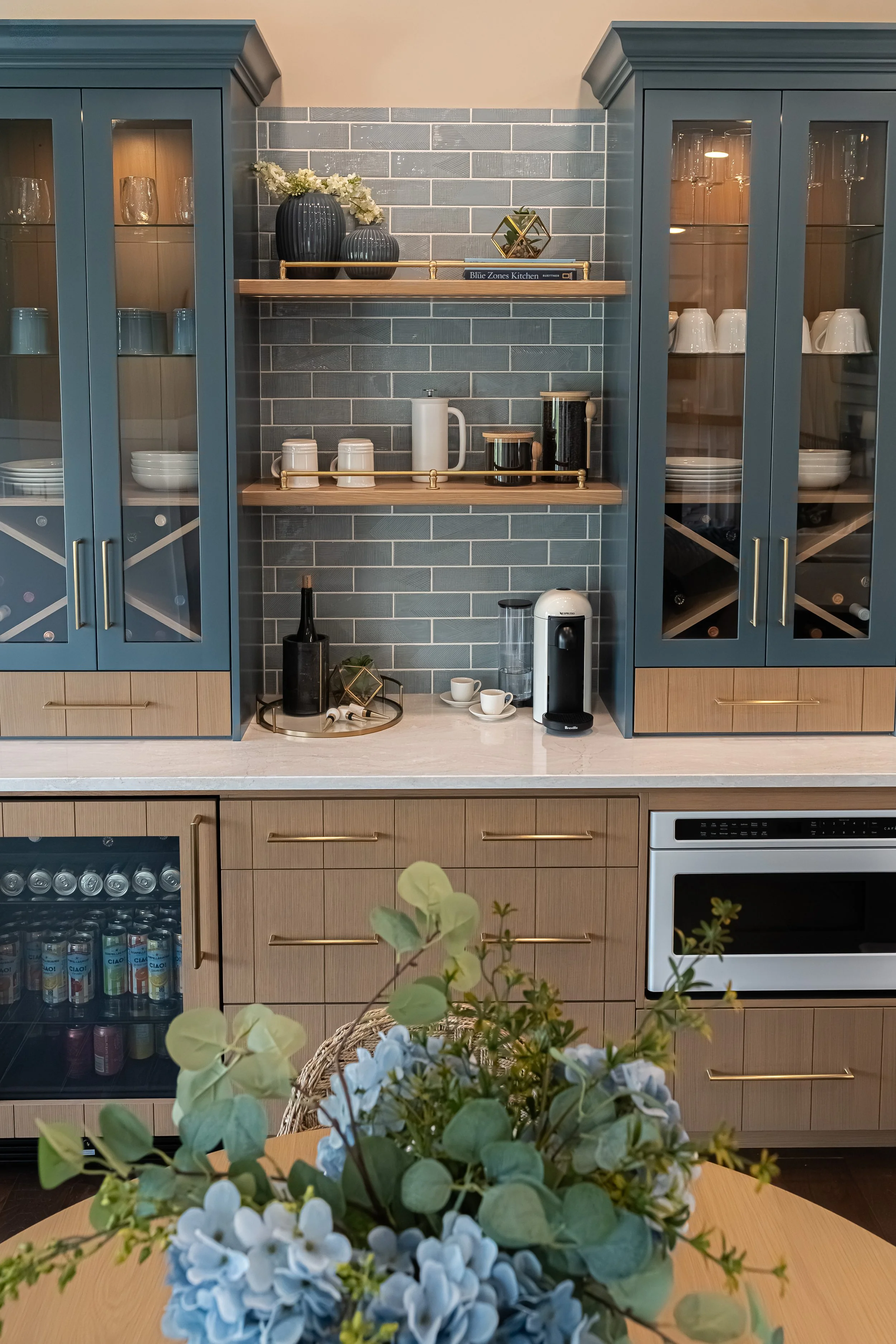 Coffee bar detail with navy glass-front cabinets, an open wood shelf styled with mugs and dishware, and gray subway tile in a Centerville home renovation.