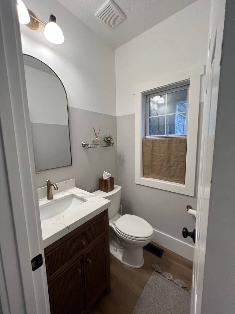 Renovated half bath with an arched mirror, wood vanity, quartz countertop, two-tone wall with picture-frame molding, and a gold sconce in a Springboro, Ohio home