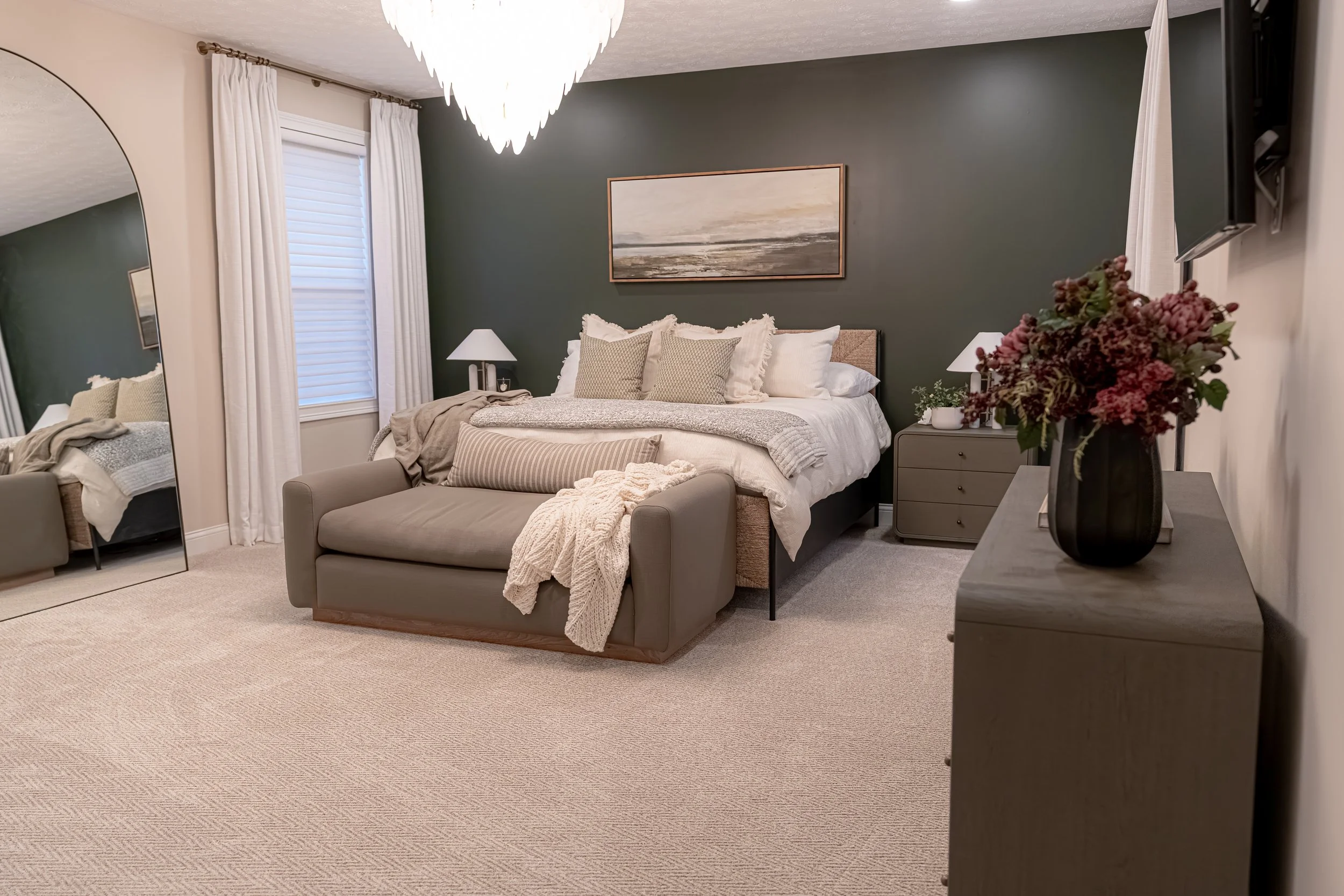 Master bedroom retreat with a deep green accent wall, layered neutral bedding, upholstered bench, and a sculptural chandelier in an Oakwood, Ohio renovation