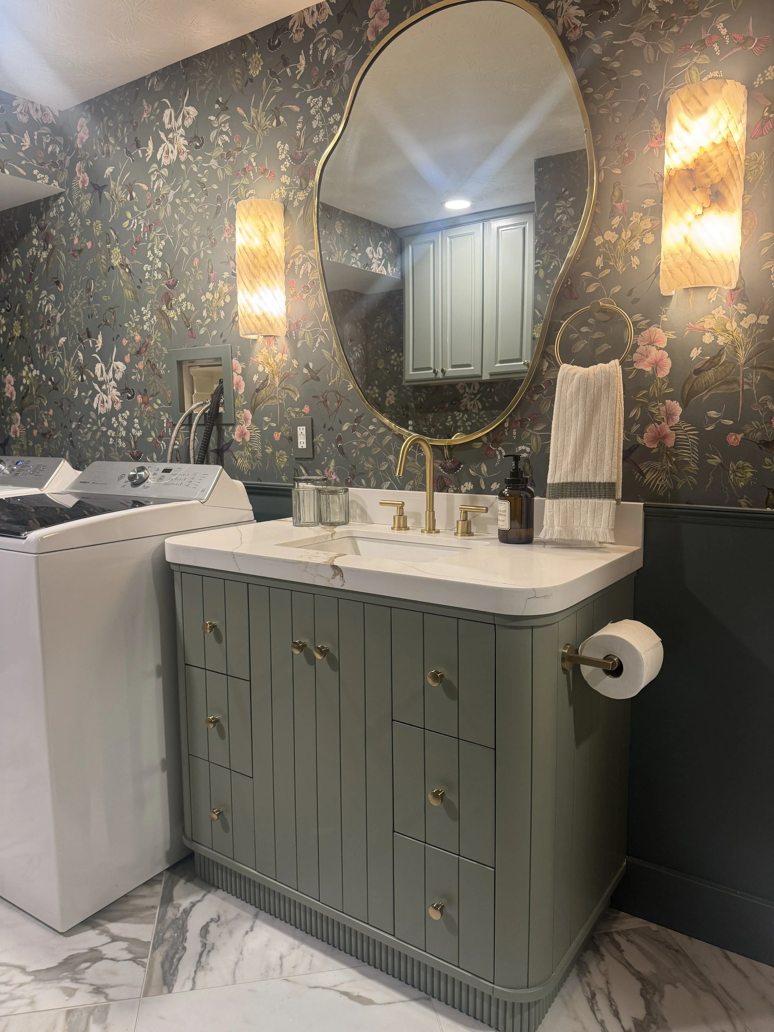 Beavercreek, Ohio basement bathroom and laundry renovation with a fluted sage-green vanity, marble countertop, botanical wallpaper, and brass fixtures by White Oak Interiors