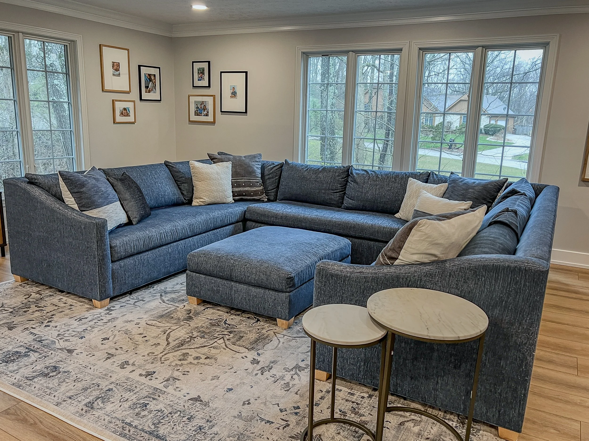 Living room with a blue sectional, brass nesting tables, and a framed gallery wall beneath a row of windows in a Centerville whole-home remodel.