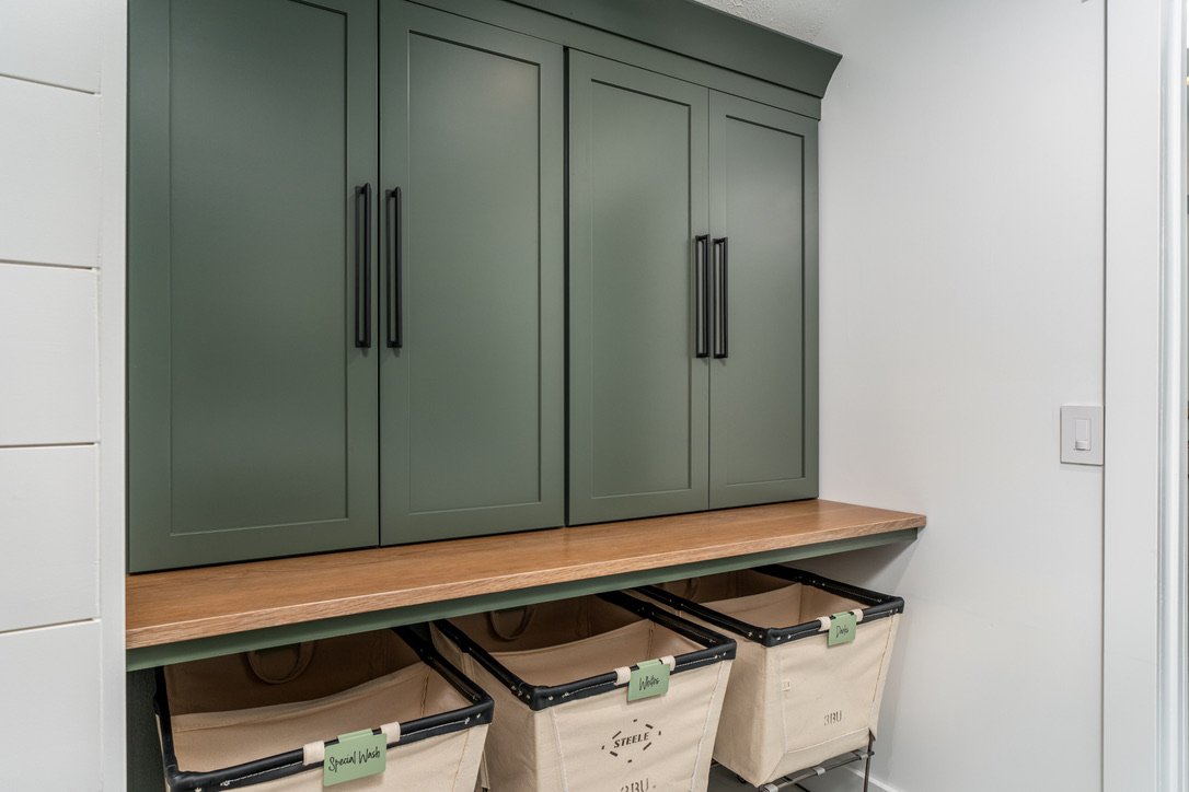 Renovated laundry room with sage green shaker cabinets, black bar pulls, a butcher-block bench, and labeled canvas sorting bins