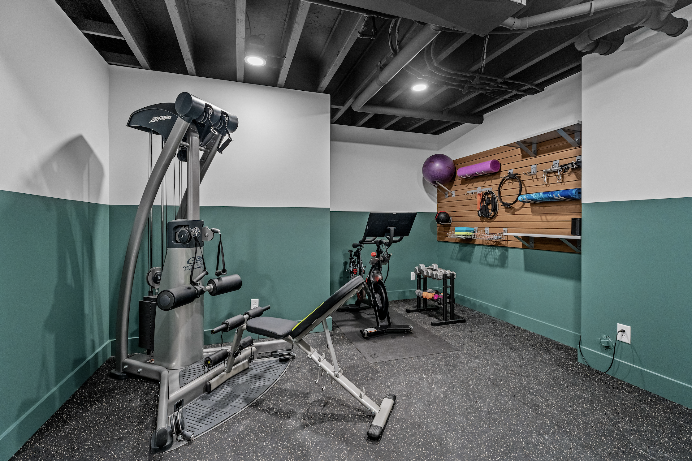 Finished basement home gym with two-tone green walls, exposed black ceiling, recessed lighting, and a wood slat wall for equipment storage