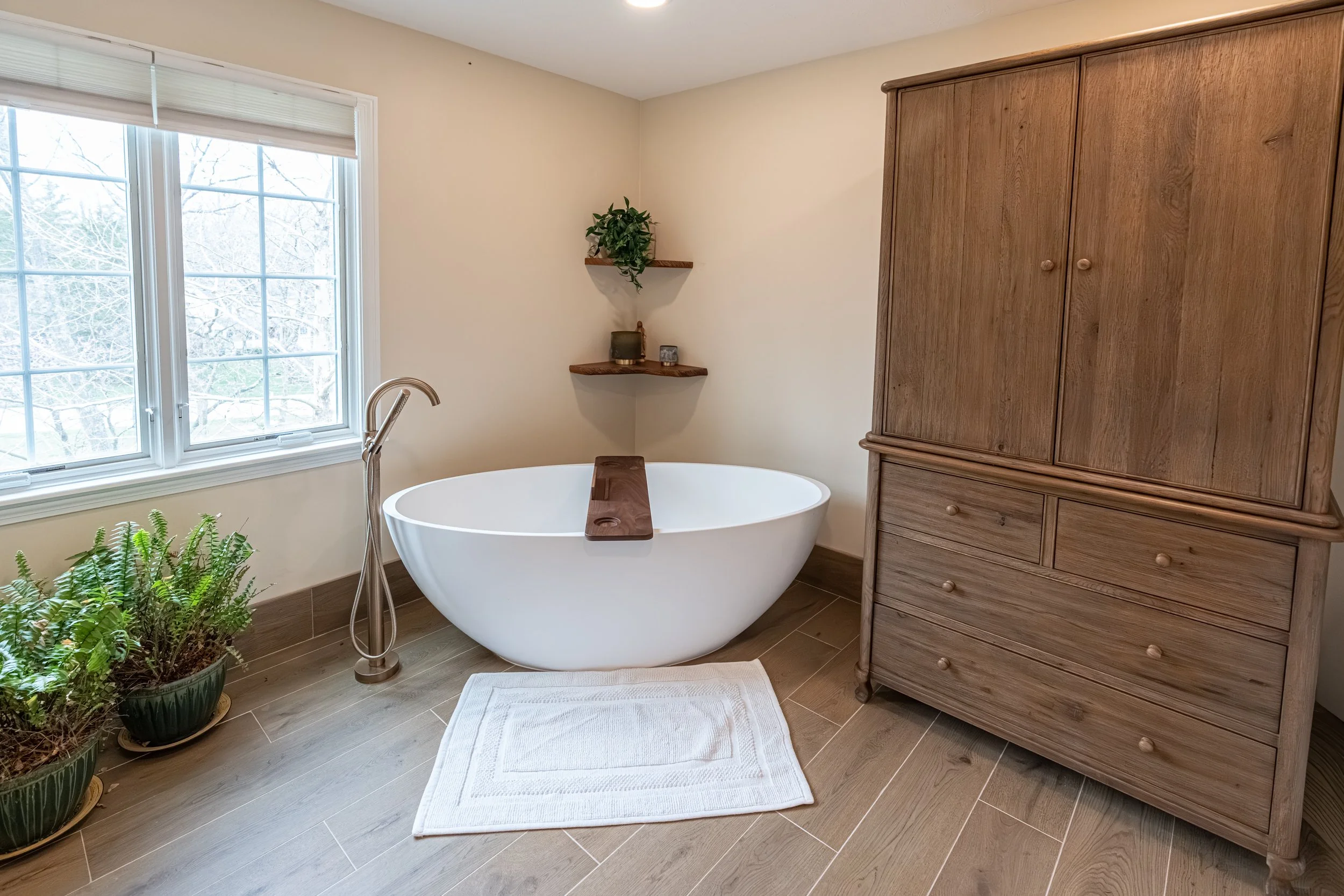 A bathroom with a white free-standing bathtub next to a large window with blinds, a potted plant below the window, two floating shelves with decorative items, a wooden cabinet, a white bath mat, and wooden floor tiles.