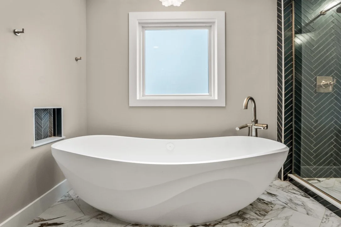 Freestanding volcanic-stone soaking tub beneath a new window with a floor-mounted gold faucet and green herringbone tile in a Xenia, Ohio master bathroom