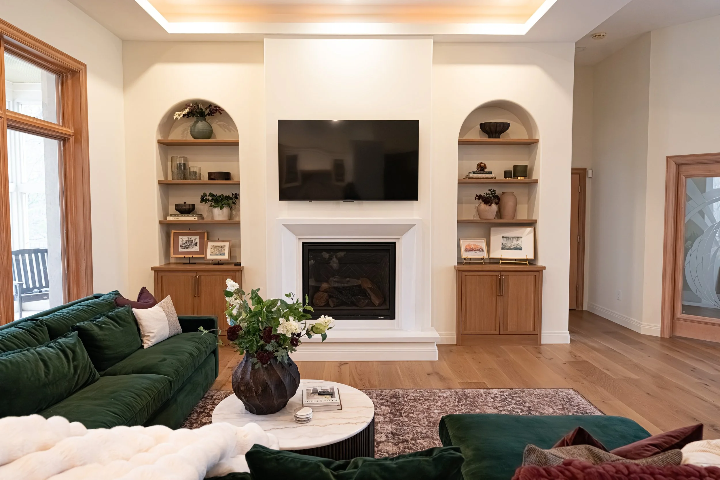 White fireplace with a mounted TV flanked by arched built-in niches with custom wood shelving in a Bellbrook living room remodel.