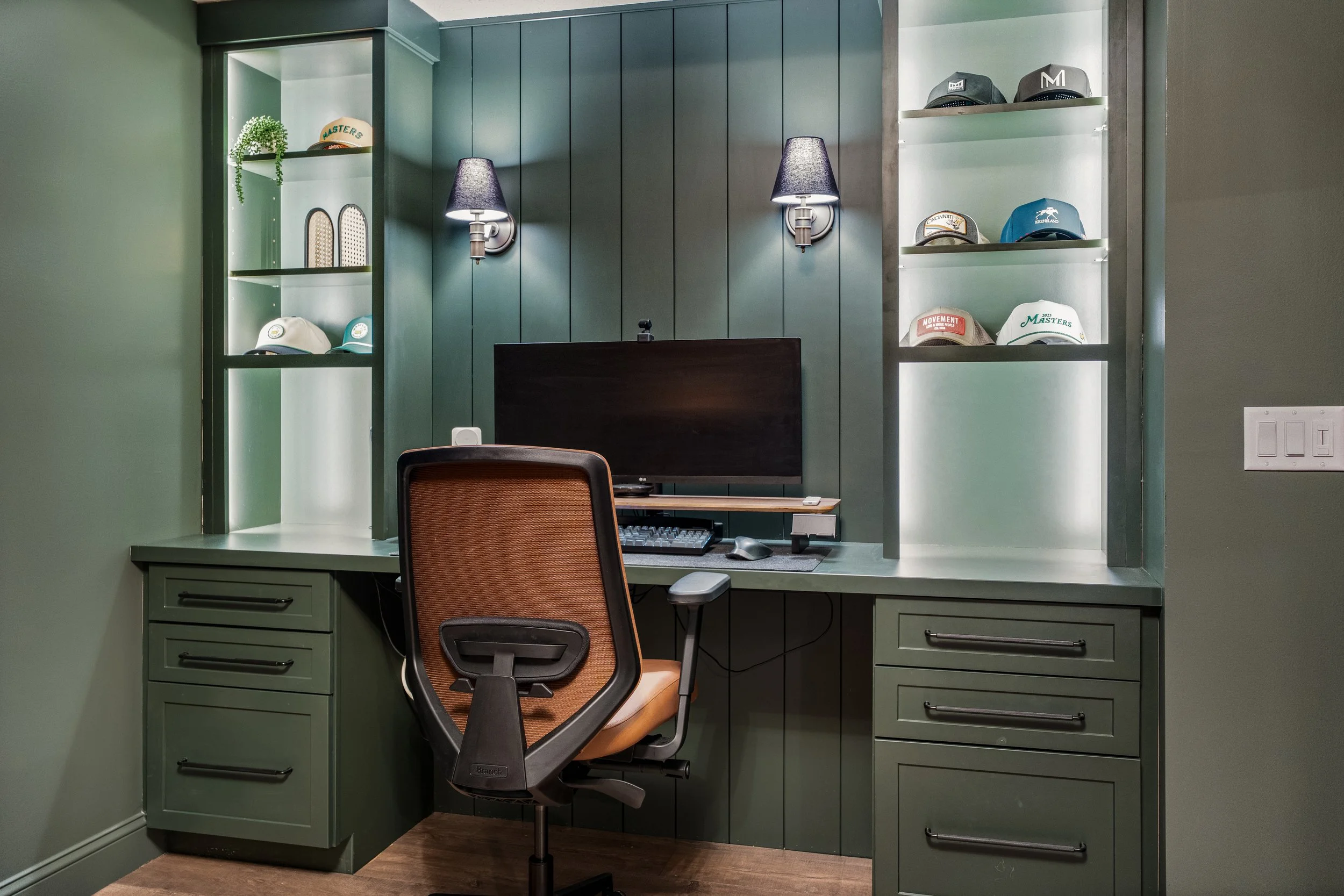 A computer workspace with green cabinetry, a desk, an office chair, and built-in shelves displaying various hats and decorative items.