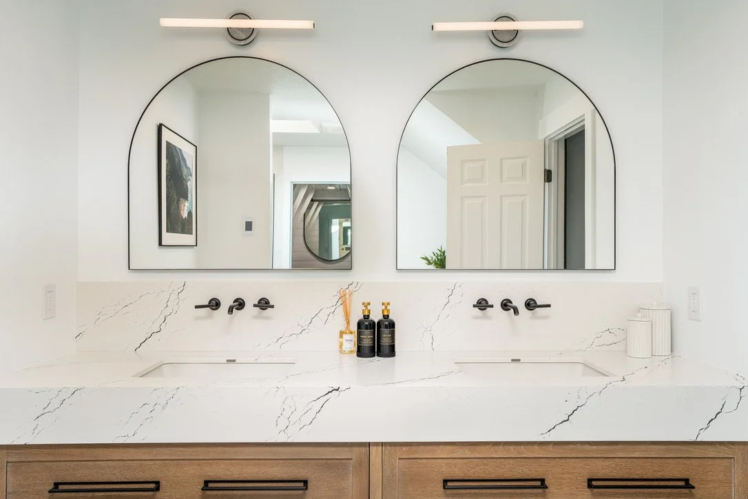 Renovated master bathroom with double vanity, arched black-framed mirrors, white oak cabinets, and white quartz counters with matte black fixtures