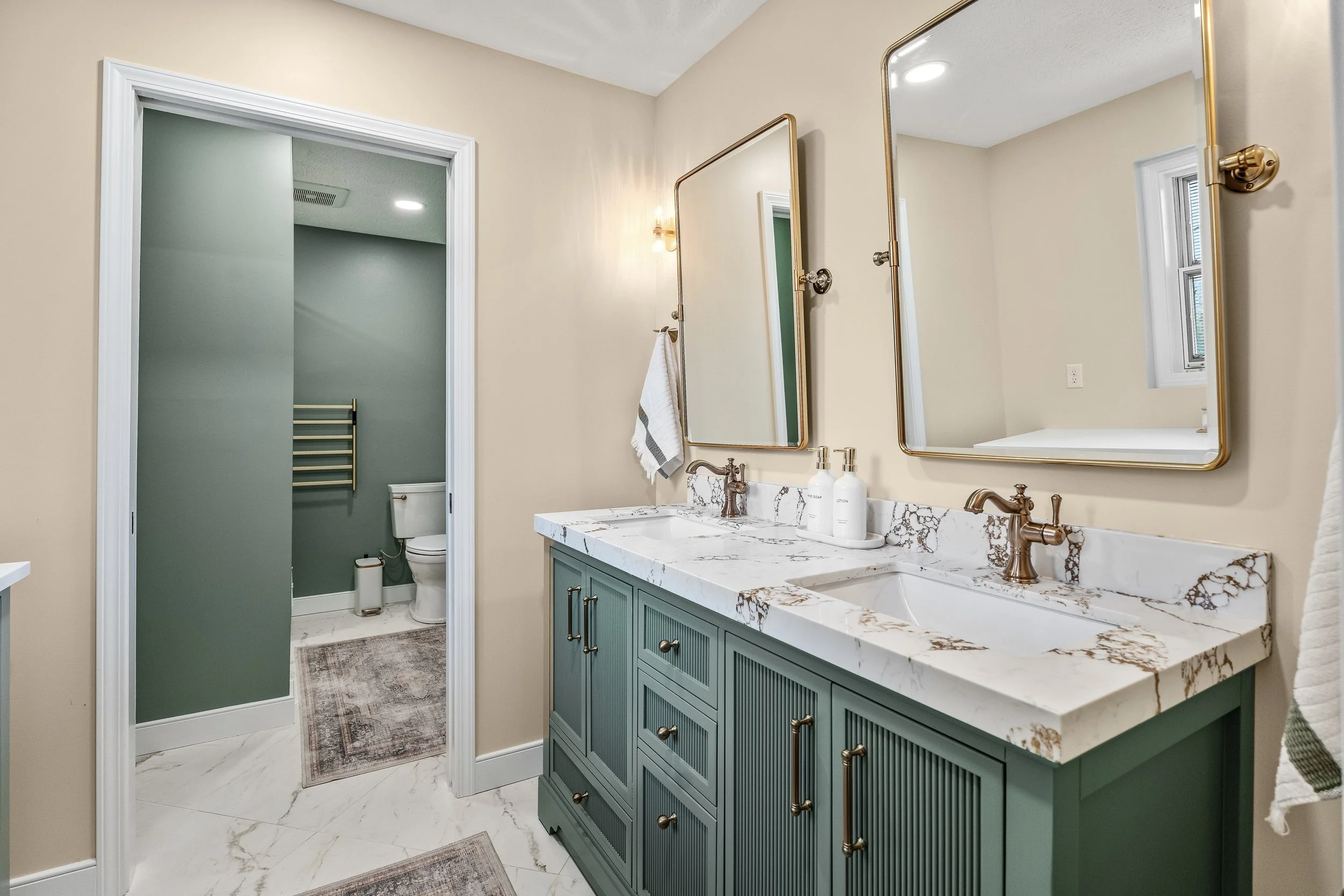 Basement bathroom vanity in Centerville, Ohio with green double vanity, gold fixtures, marble countertop, and view to separate shower room by White Oak Interiors