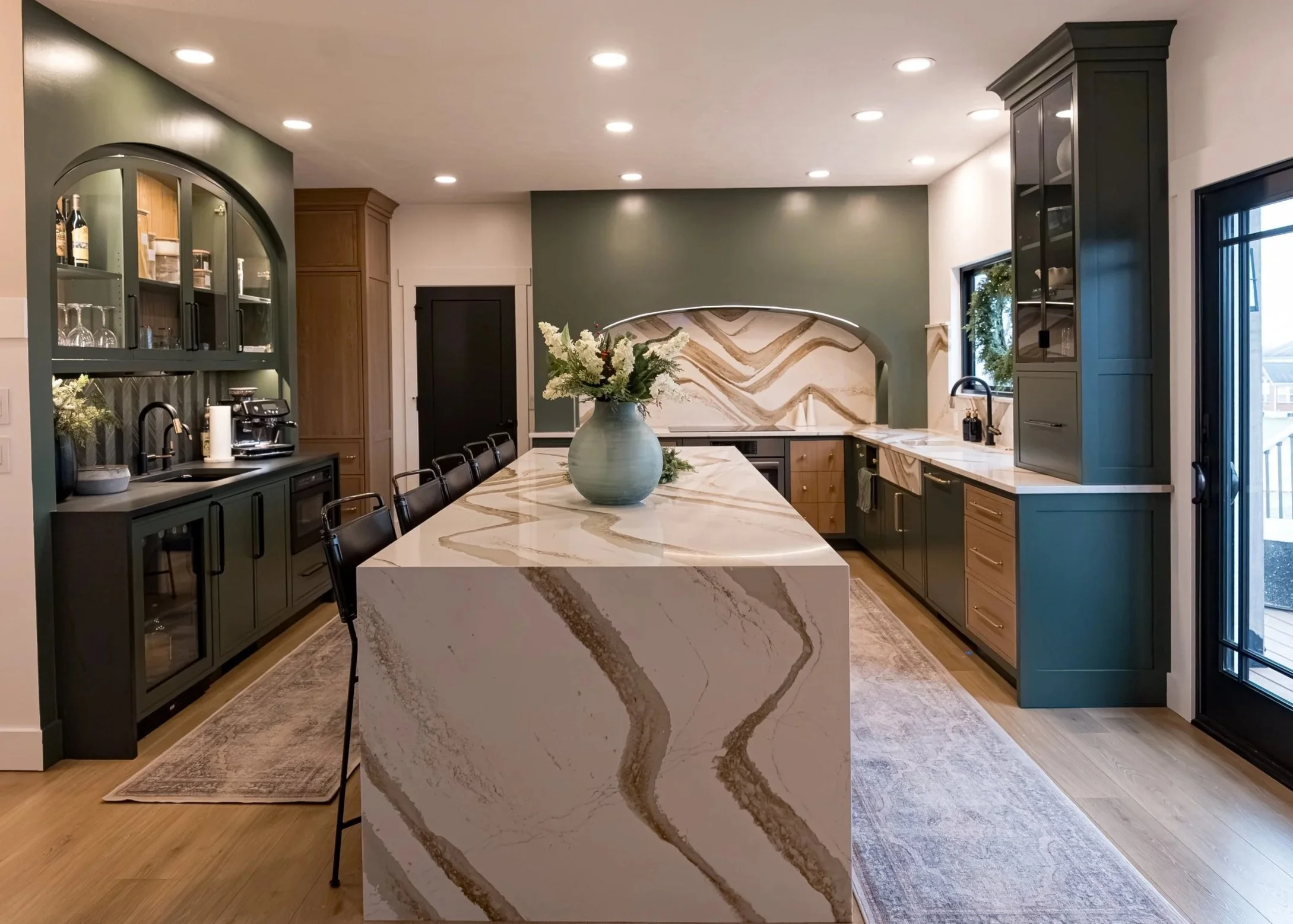 Modern kitchen with green and wooden cabinetry, marble island with a vase of flowers, built-in appliances, and large windows with black frames.