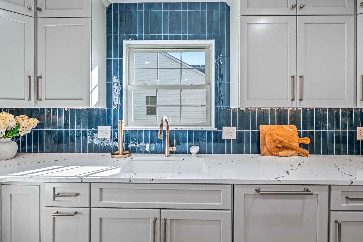 Kitchen with white cabinets, marble countertop, blue tiled backsplash, window above sink, and a cutting board on the counter.