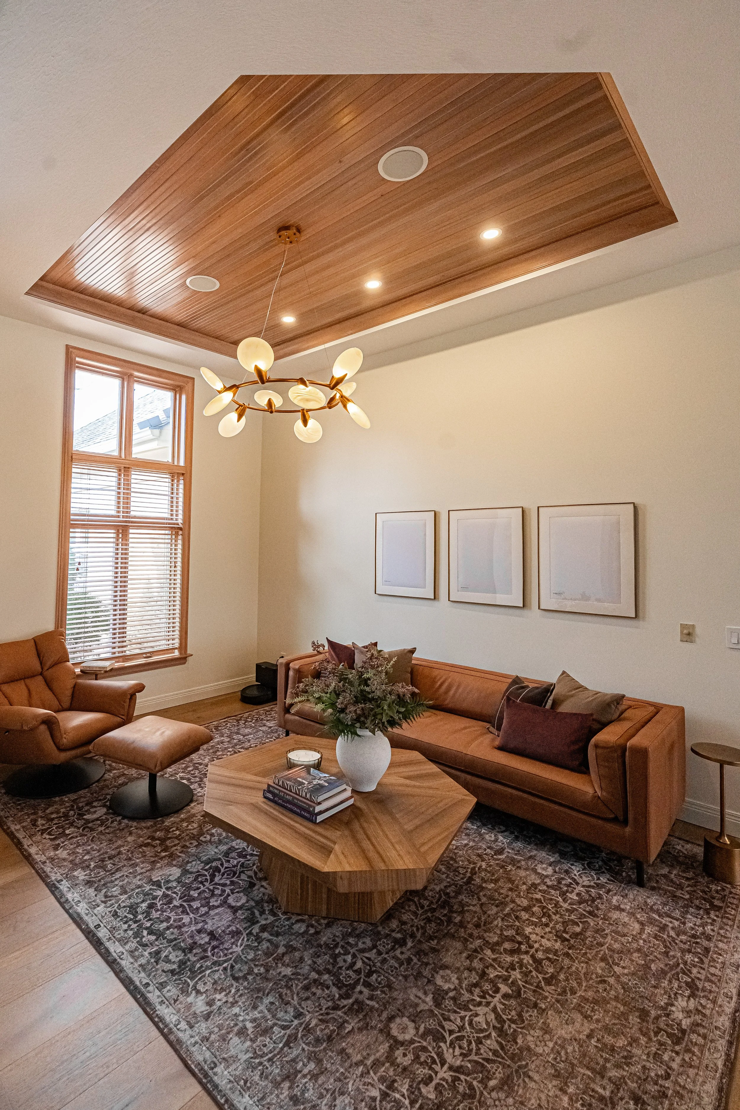 Lounge room with a wood-slatted tray ceiling, modern chandelier, and tan leather sofa beside a tall wood-trimmed window in a Bellbrook whole-home remodel.
