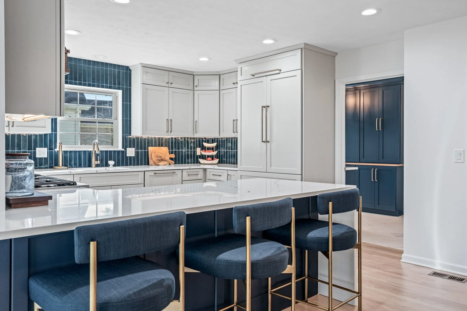 White and navy kitchen with a quartz peninsula, blue-and-brass stools, and blue tile backsplash in a Beavercreek, Ohio remodel.