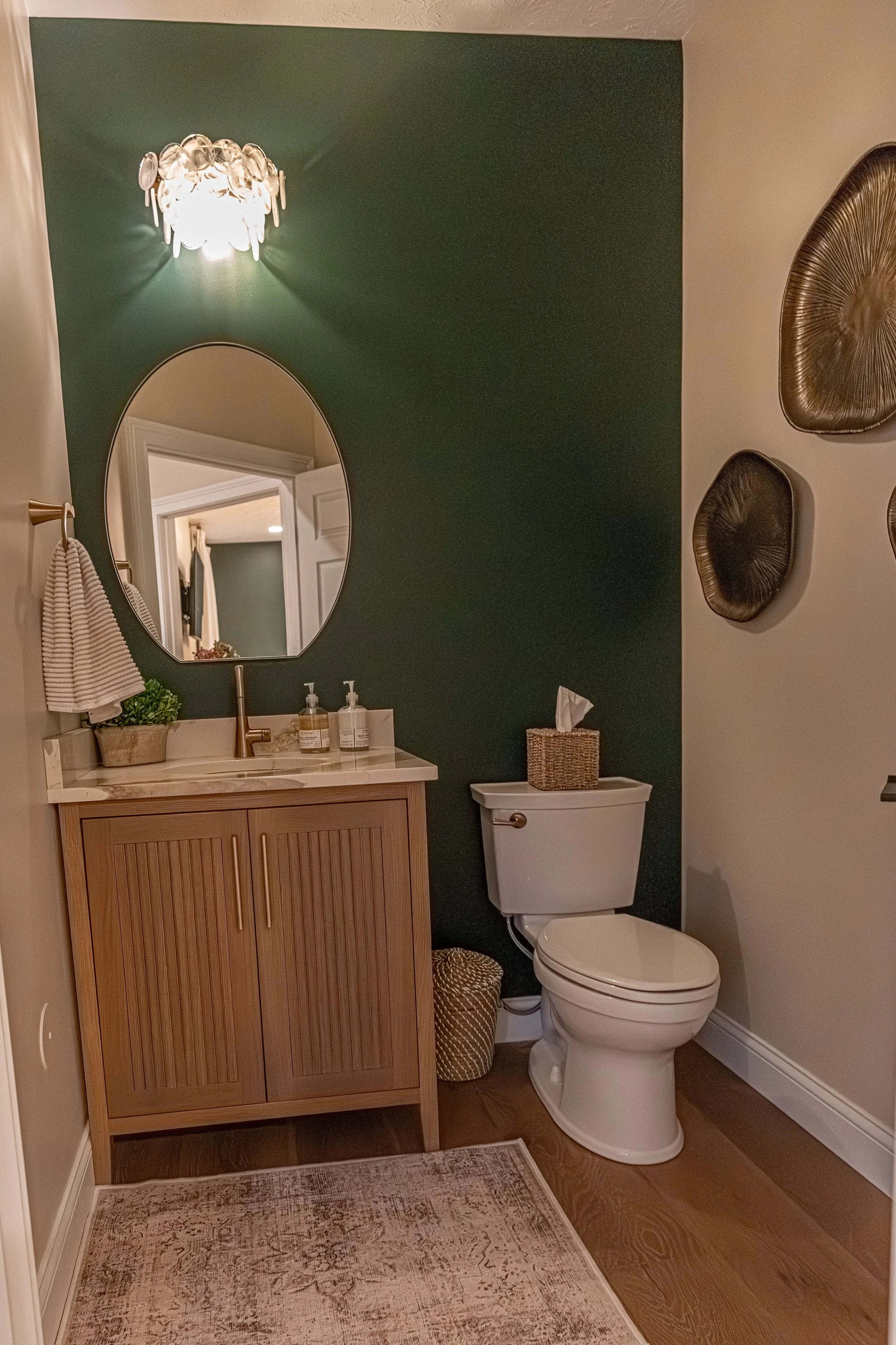 Half bath with a deep green accent wall, custom reeded inset vanity, egg-shaped flat-bottom mirror, gold light fixture, and woven wall art in an Oakwood, Ohio home