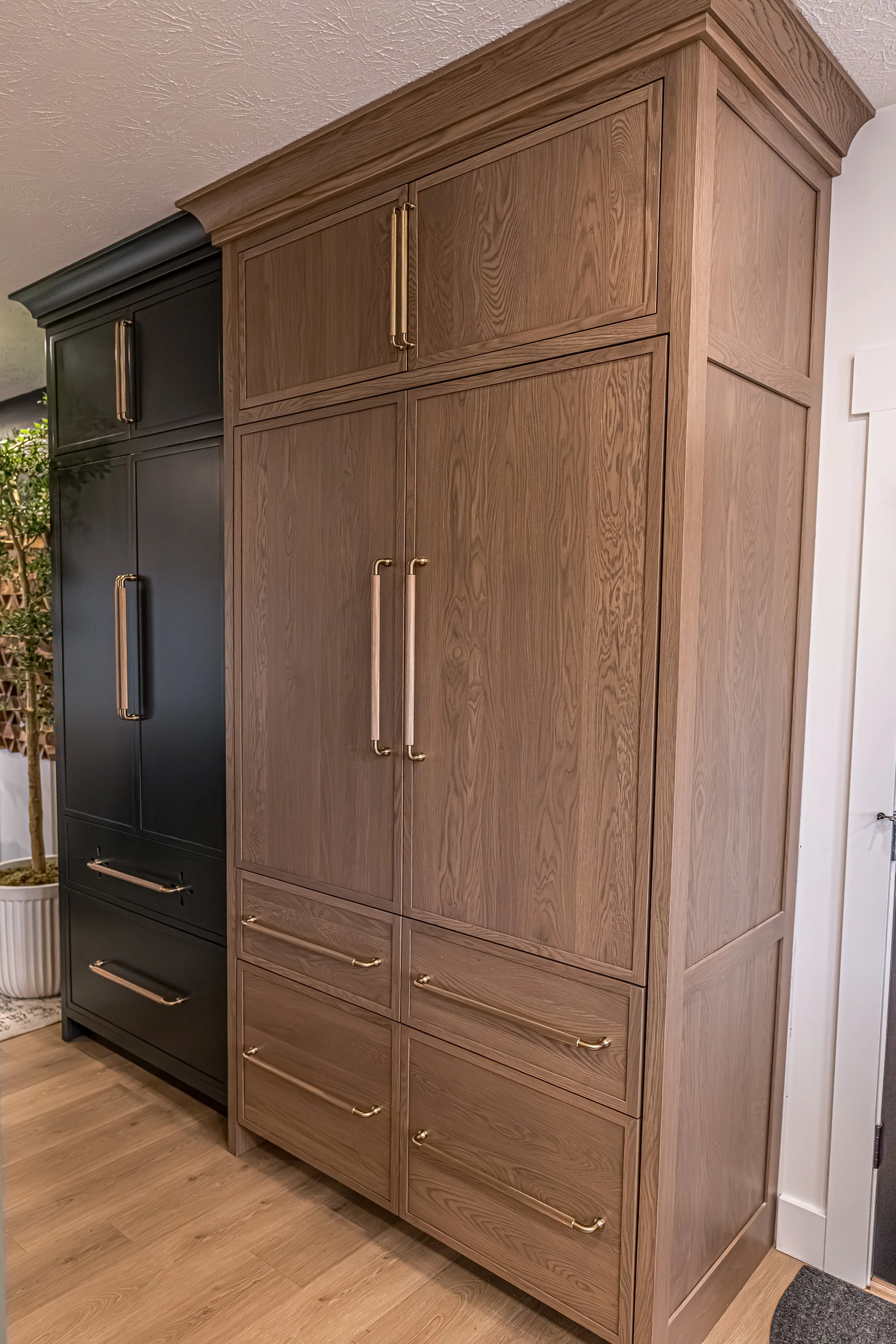 Floor-to-ceiling white oak pantry cabinets with brass hardware beside a black paneled cabinet