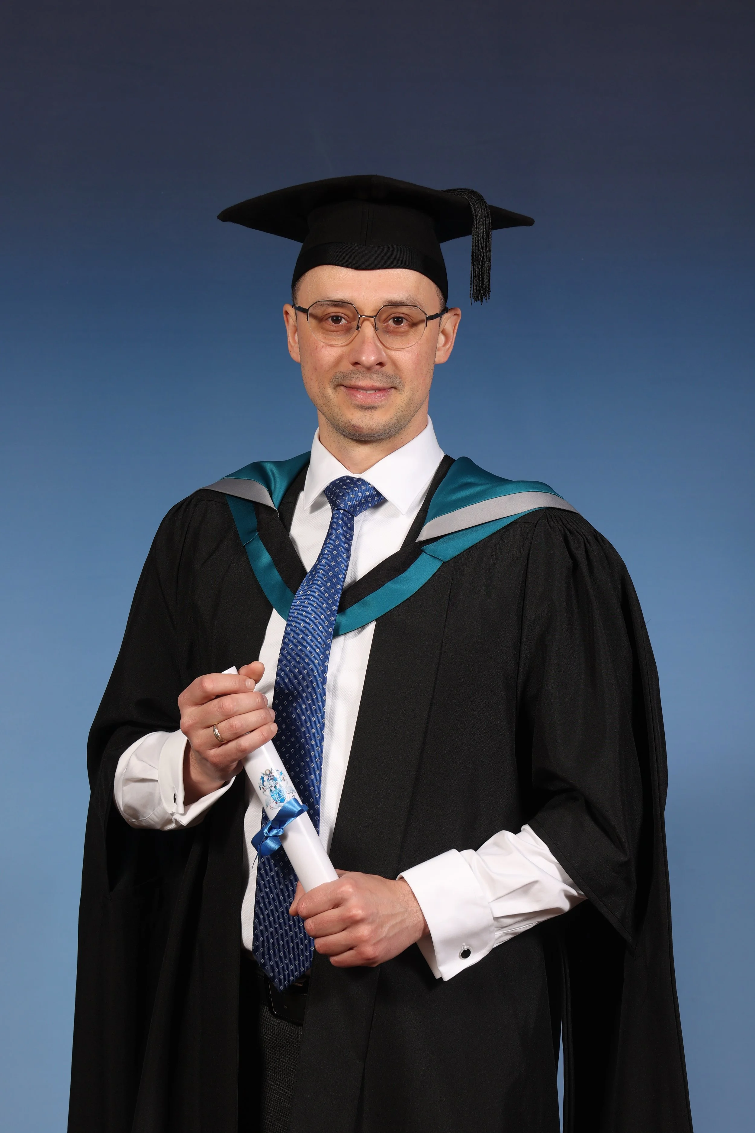 A man wearing glasses in a graduation gown and cap, holding a diploma against a blue background.
