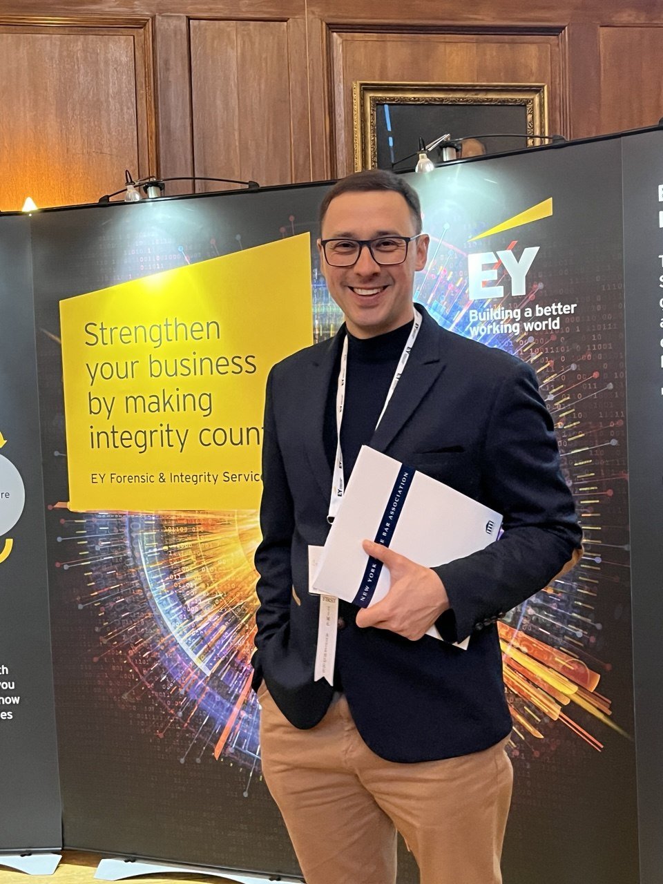 A man wearing glasses, a black blazer, and khaki pants standing in front of an EY conference booth holding a folder, smiling at the camera.