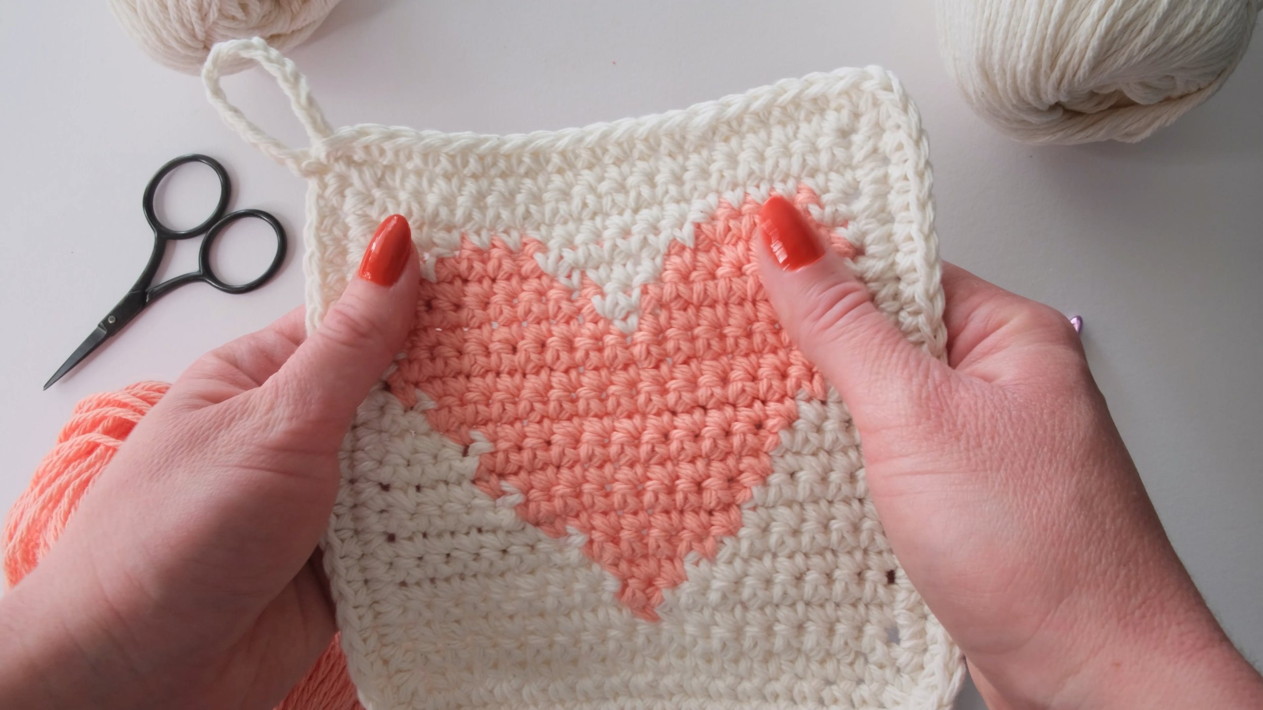 Hands holding a crocheted square potholder with a pink heart in the center, surrounded by cream-colored yarn and crochet scissors on a light background.