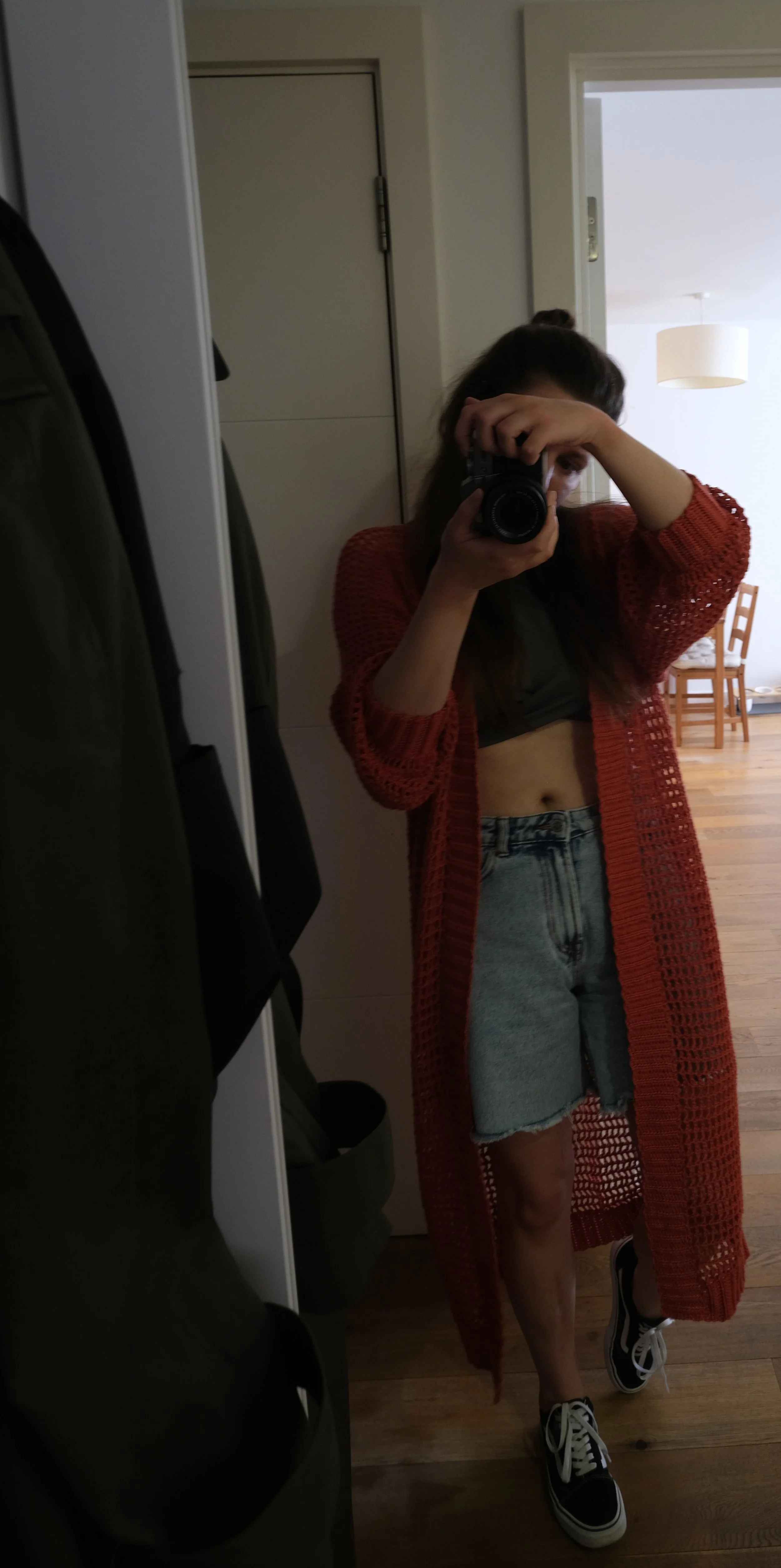A young woman is taking a mirror selfie in a hallway, holding a camera in front of her face. She is wearing a black crop top, a red open-knit cardigan, denim shorts, and black and white Vans sneakers.