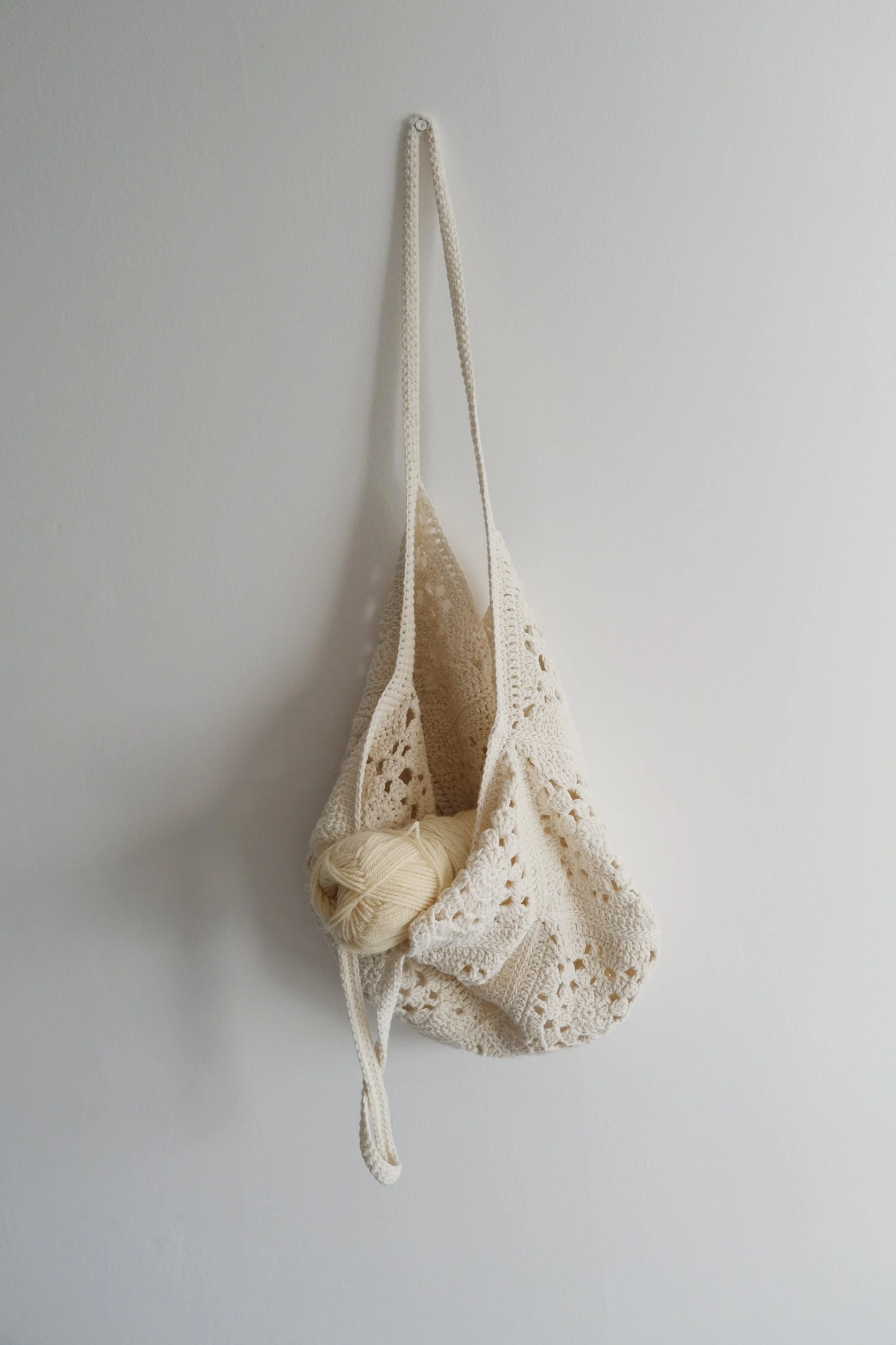 A cream-colored crocheted bag with a ball of yarn inside hanging on a plain white wall.