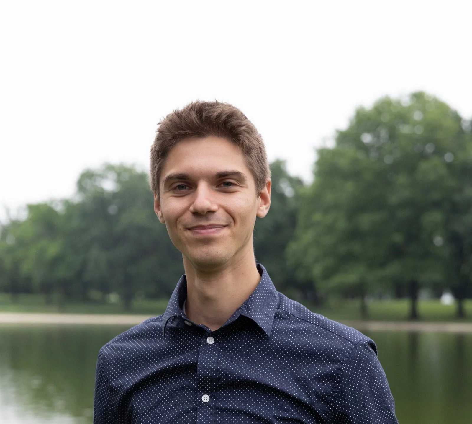A young man with short brown hair and a slight smile, wearing a navy blue, polka-dotted button-up shirt, standing outdoors near a body of water with trees in the background.