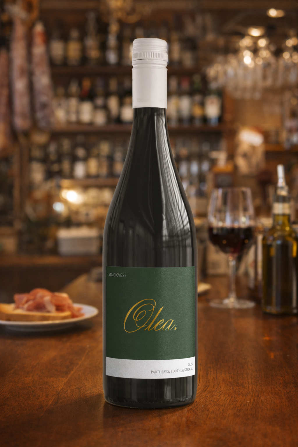 A dark wine bottle with a green label reading 'Olea' in gold cursive, placed on a wooden bar counter with a glass of red wine, a plate of prosciutto, and a blurred background of bar shelves and glasses.