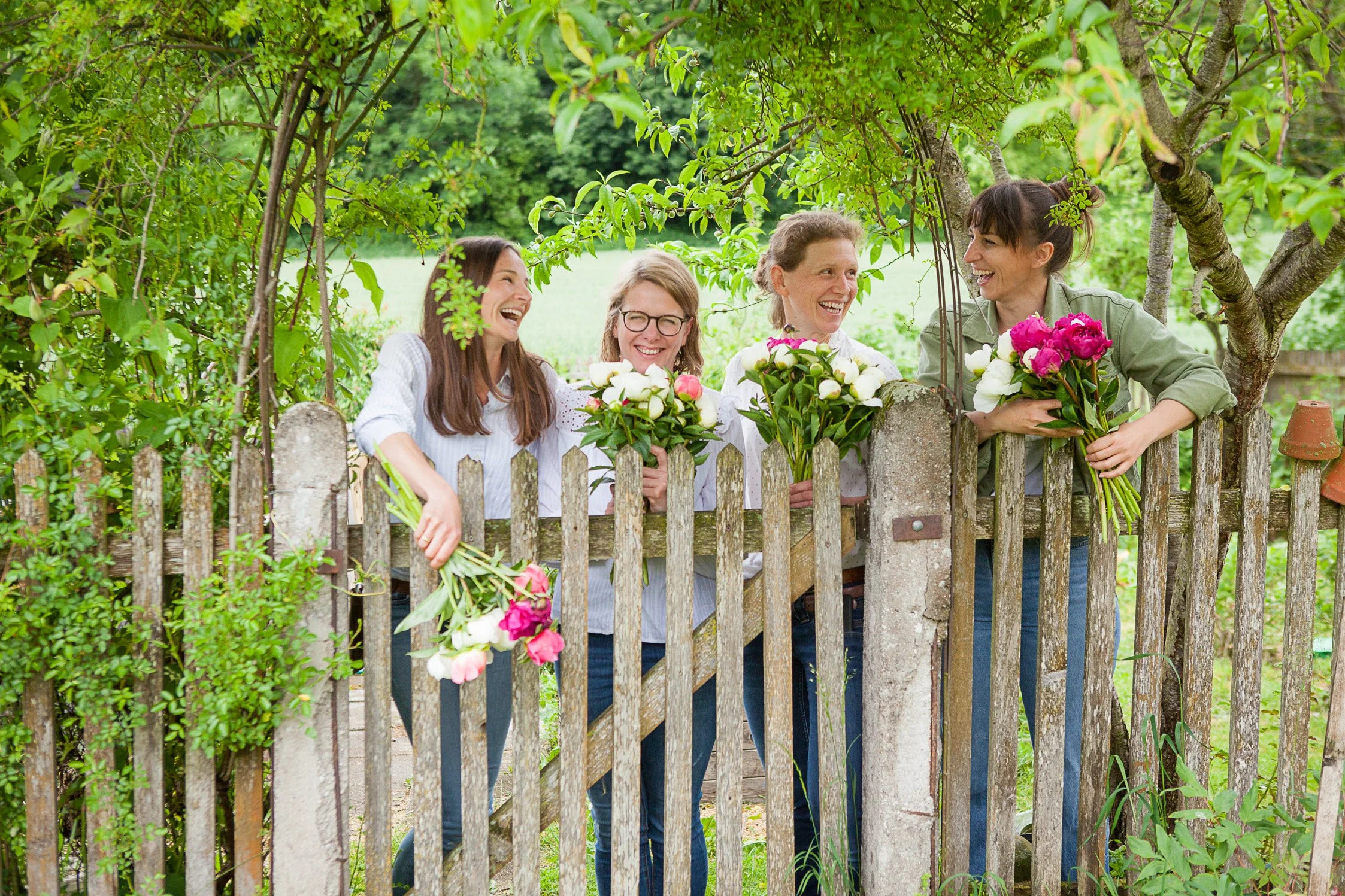 Fünf lachende Frauen mit Blumen stehen hinter einem Zaun unter einem Baum in einem Garten.