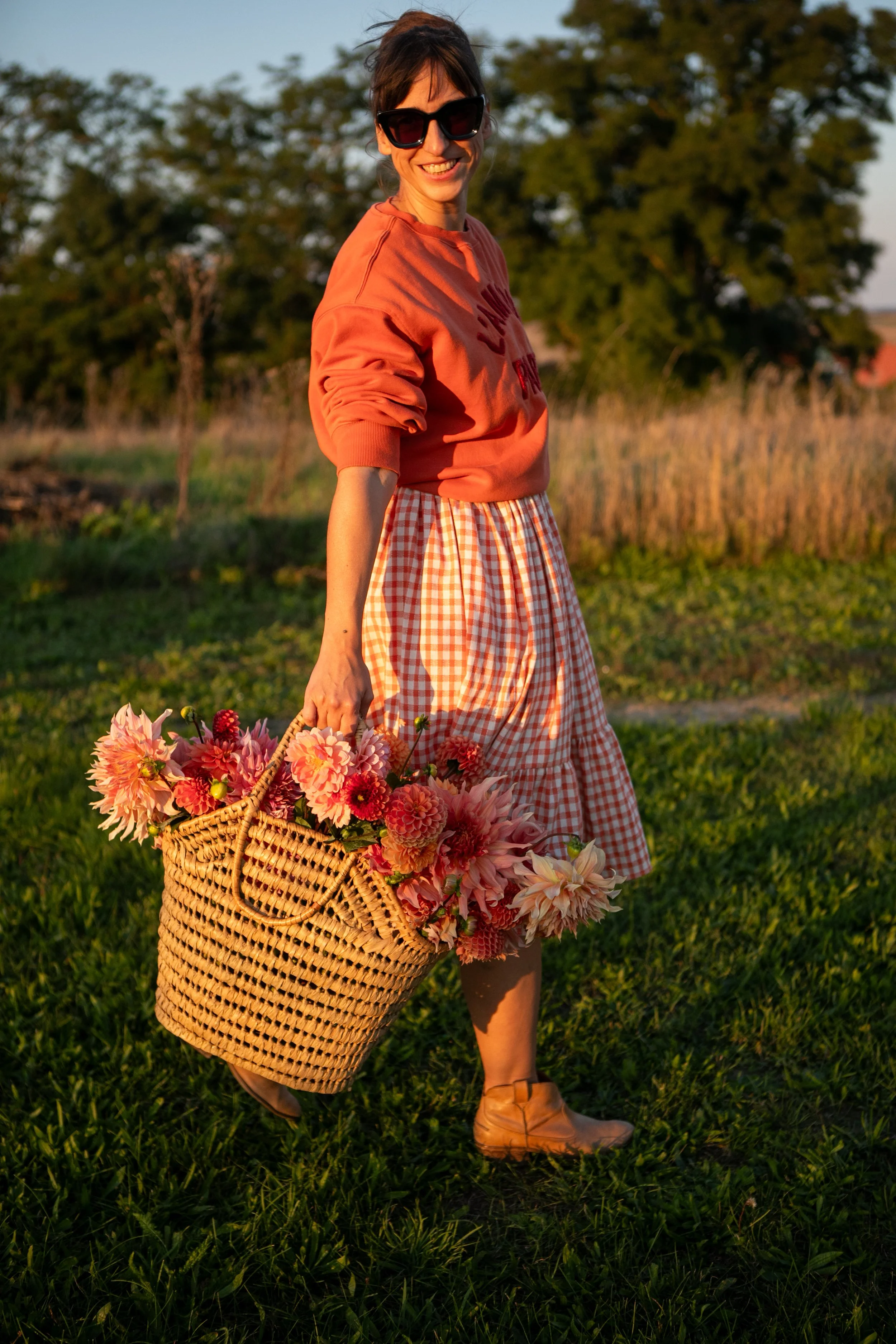 Frau trägt orange Pullover und Tuch, hält Korb voller bunter Blumen, lacht beim Spaziergang im Grünen bei Sonnenuntergang.