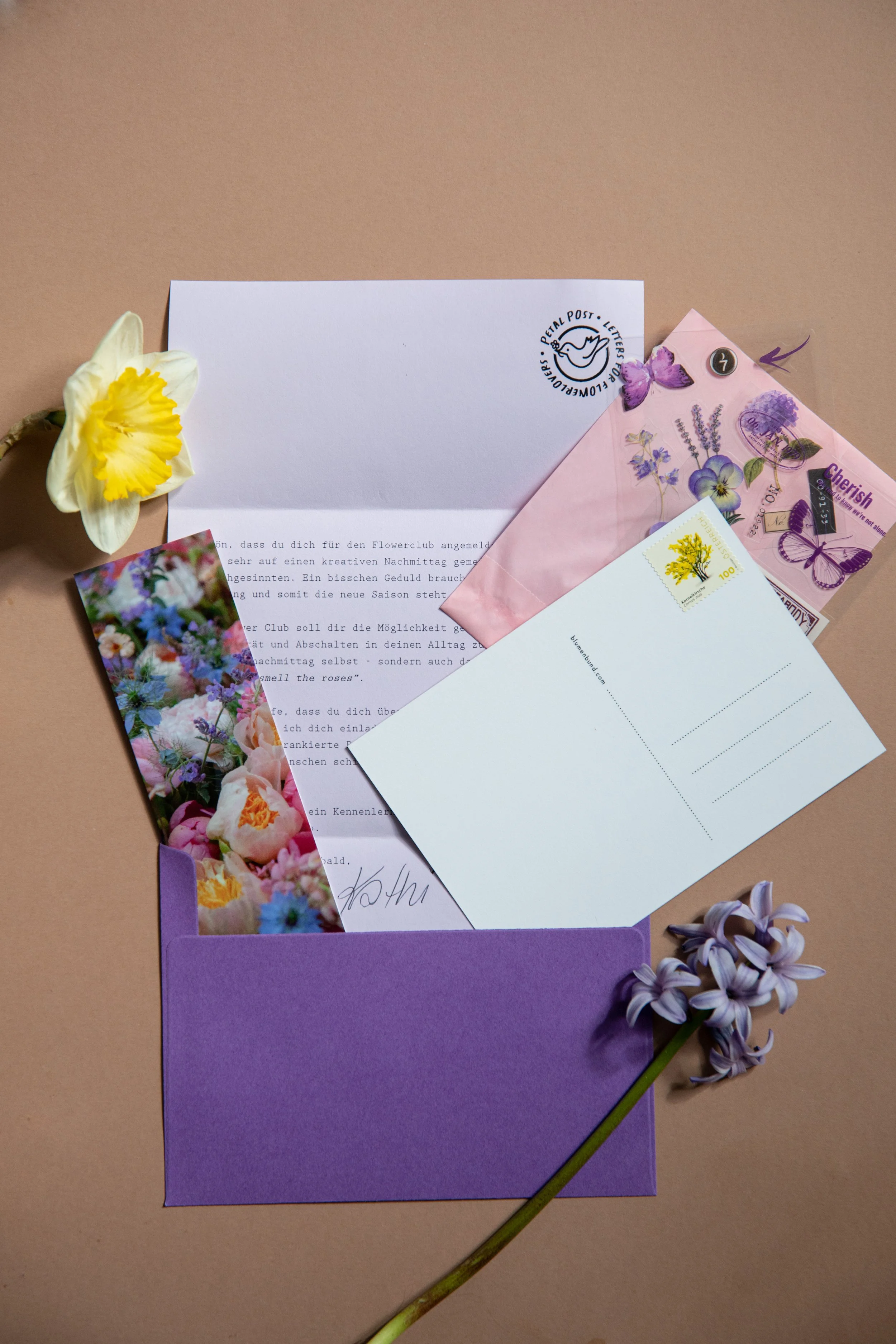 Ein gelber Narzisst mit weißen Blüten, Umschläge, Briefe, eine Postkarte mit buntem Blumendesign, ein lila Umschlag, eine Postkarte mit einem Blumenbild, rote, violette und lilafarbene Blumen, ein lilafarbener Briefumschlag, und blühende Lavendel- und Hyazinthenblüten auf hellbraunem Hintergrund.
