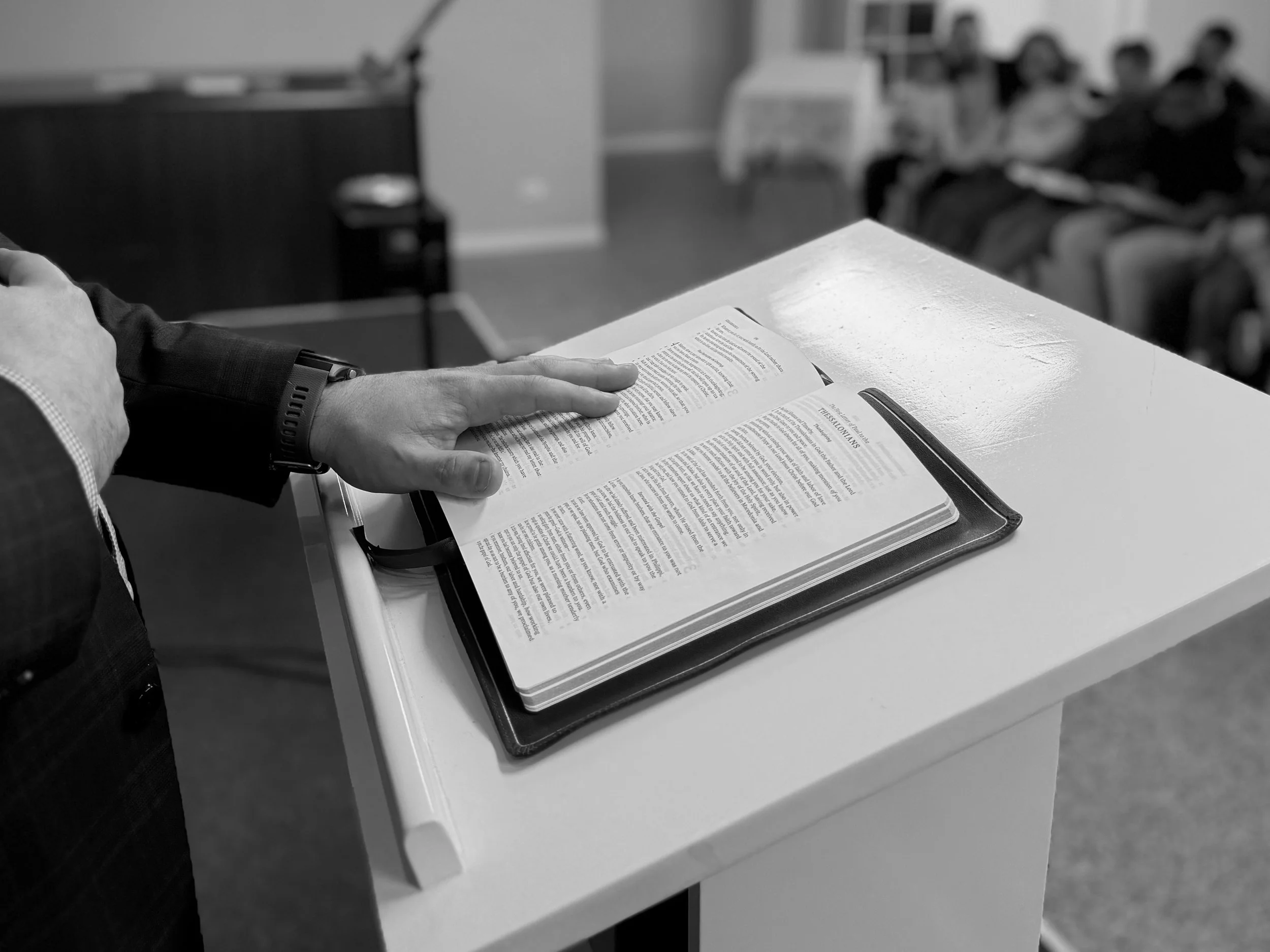 A person in a suit reading a book, standing at a lectern or podium in a room with chairs and people seated in the background.