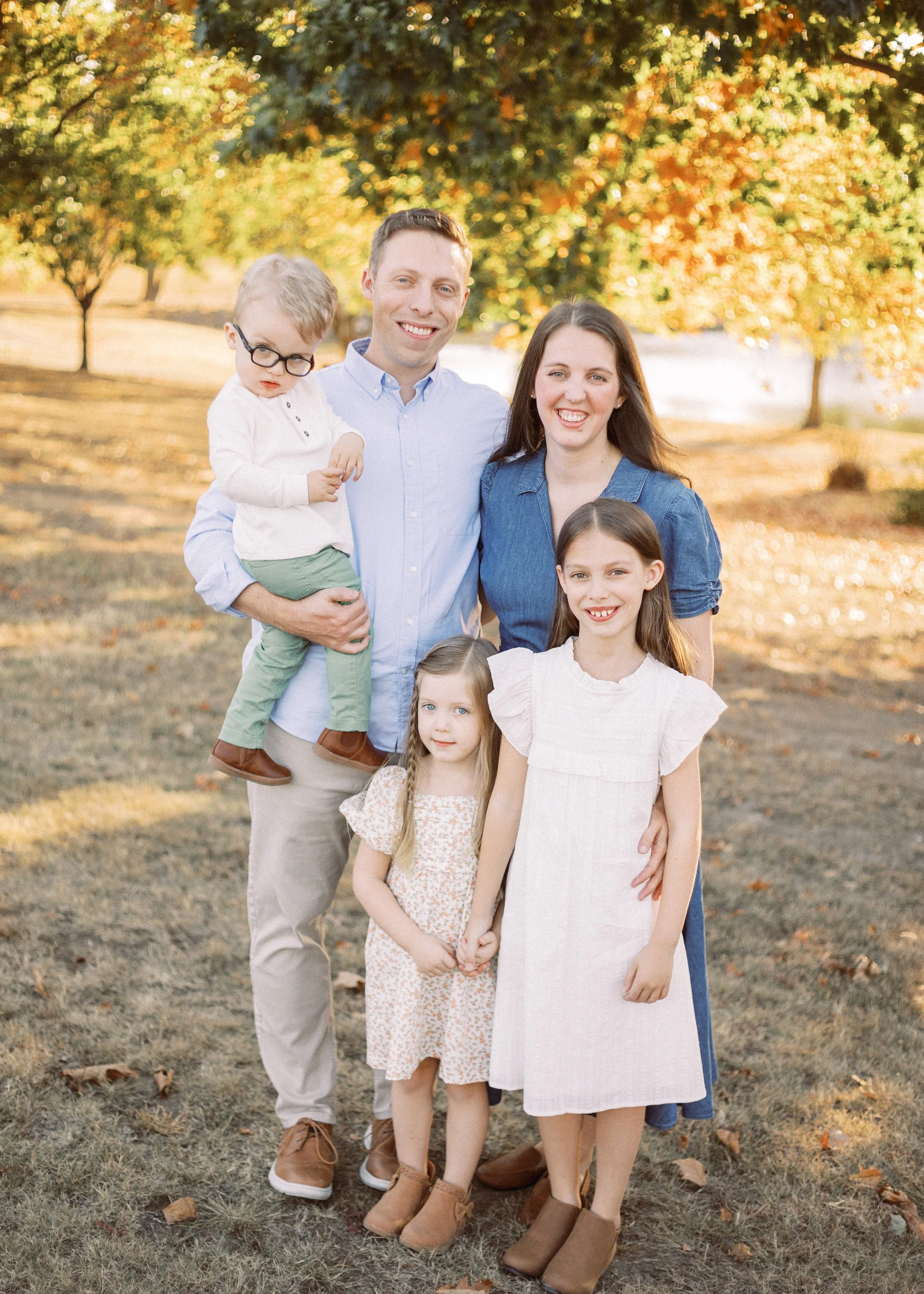 A family of six posing outdoors in an autumn park. The family includes a man, a woman, and four children, with a background of trees with fall foliage. All are smiling, dressed casually, and standing on grass with fallen leaves.