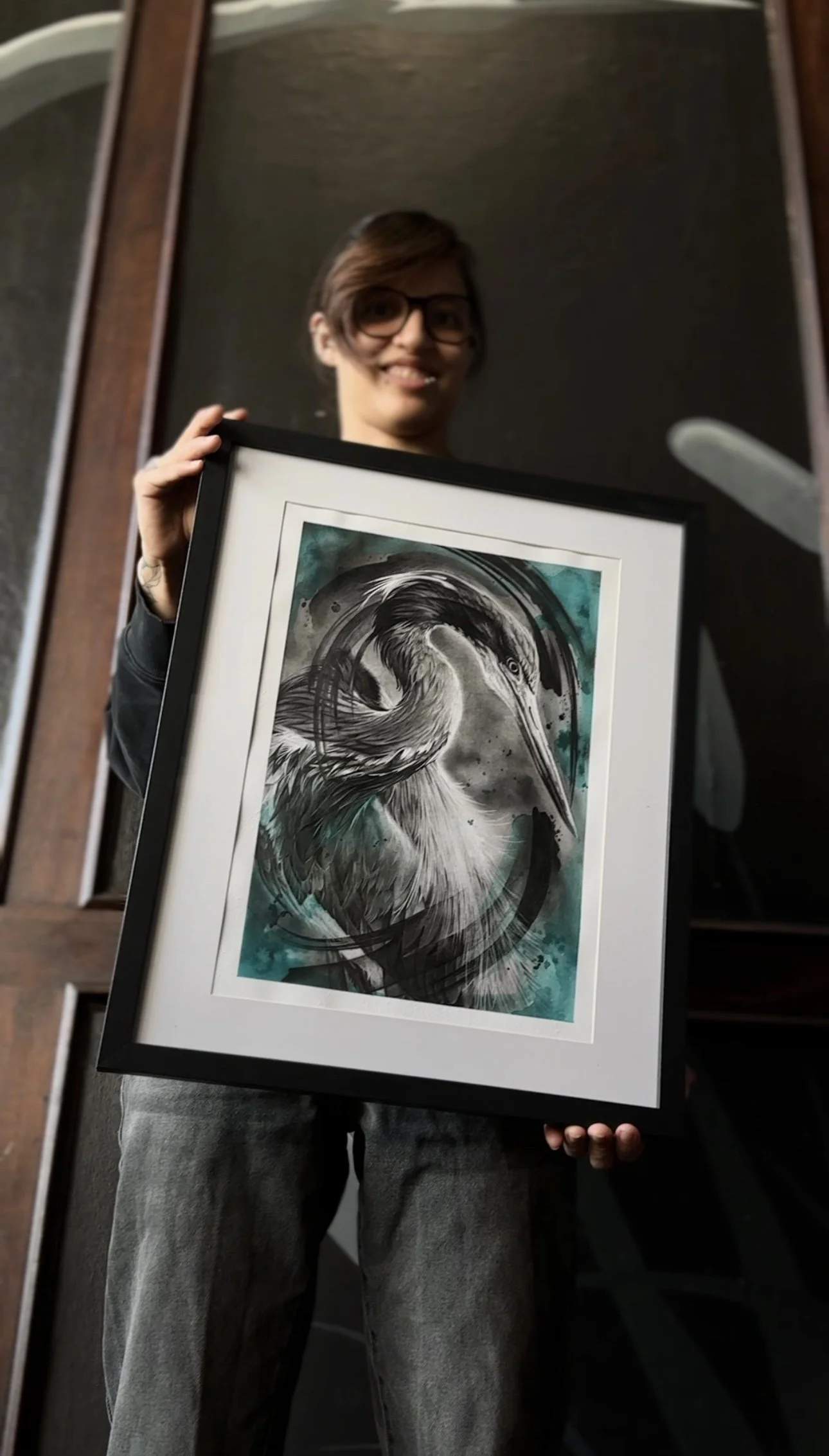 Kimmi Gayle  holding up a framed artwork of a bird,  a heron, with a watercolor-like background in shades of teal and black.