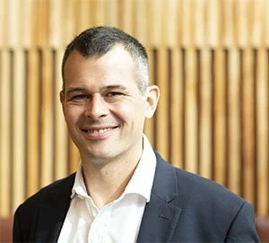 A man in a suit smiling in front of wooden paneling background.