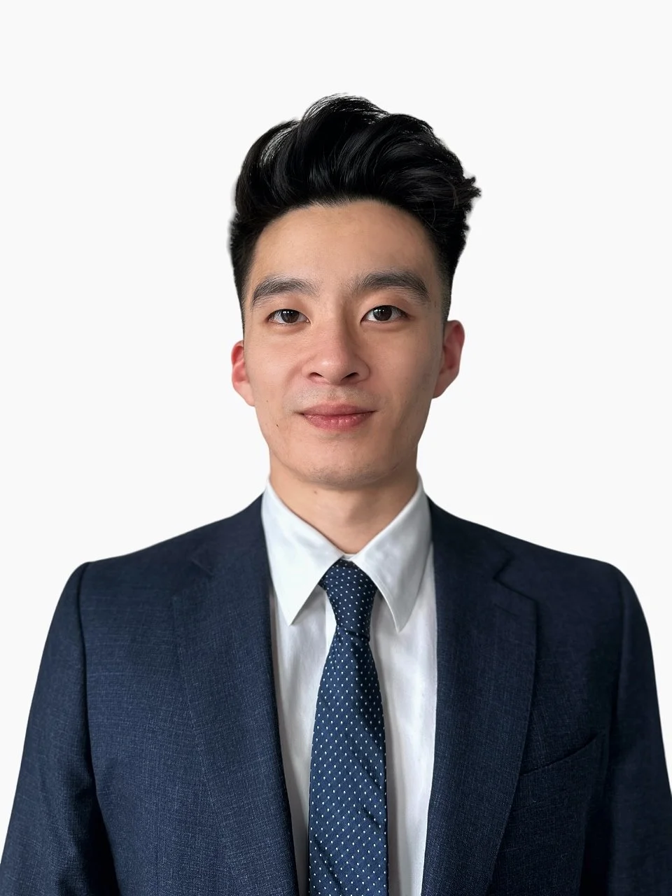 A young man with black hair and light skin, wearing a dark blue blazer, white shirt, and a blue polka dot tie, standing against a plain white background.