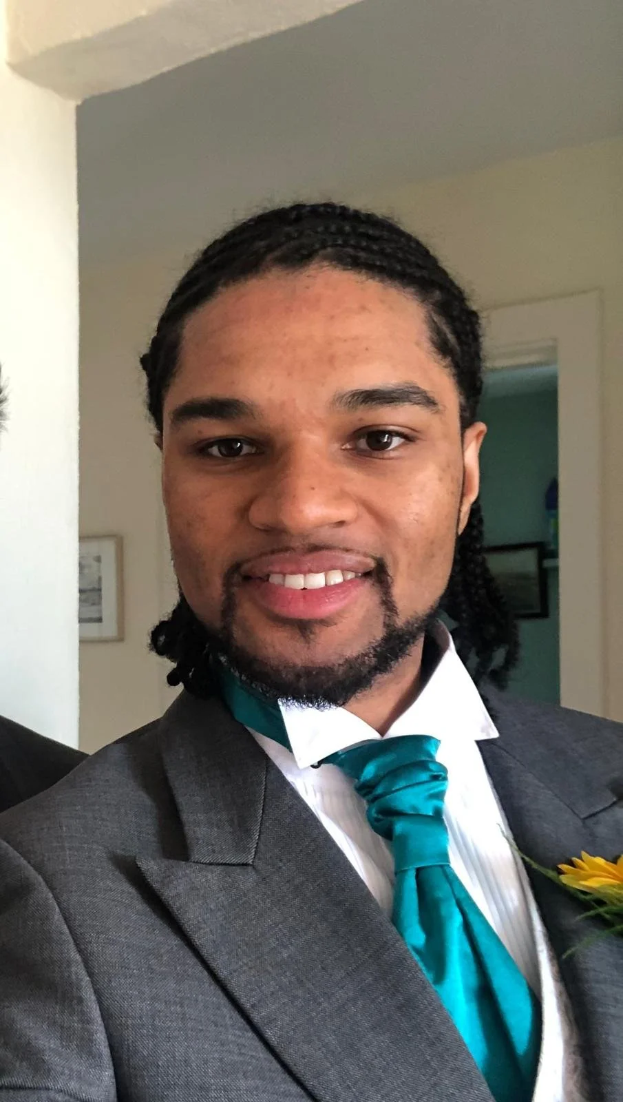 Young man with braided hair and a goatee, dressed in a formal gray suit, white shirt, turquoise tie, and a sunflower boutonniere, smiling indoors.