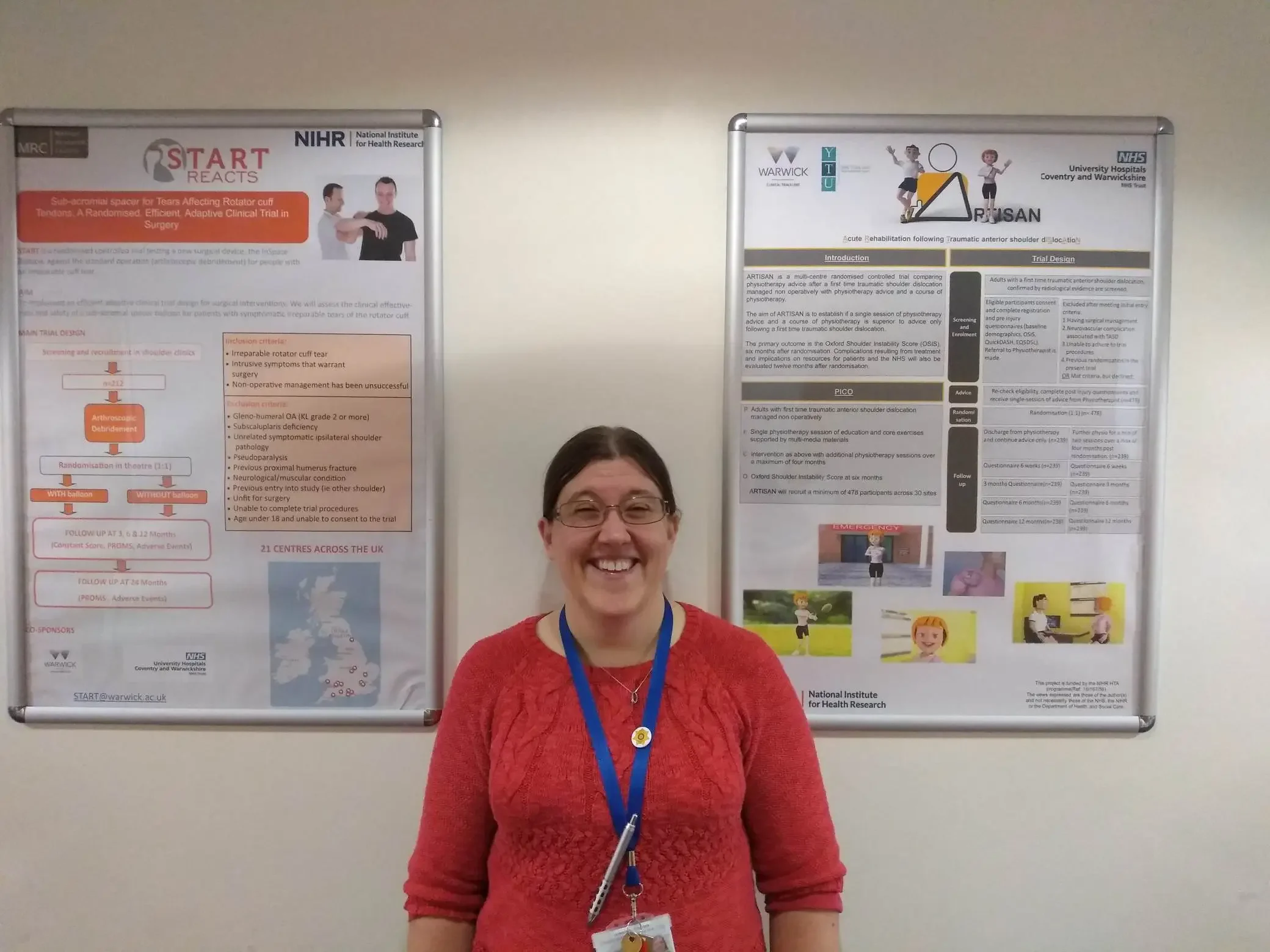 A smiling woman with glasses and a red sweater standing in front of two academic posters on a wall.