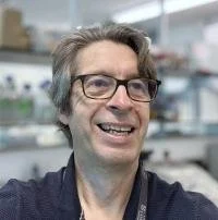 A man with gray hair and glasses smiling in a laboratory setting.