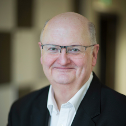 Headshot of a middle-aged man with glasses, smiling, dressed in a white shirt and dark blazer, in an indoor setting.