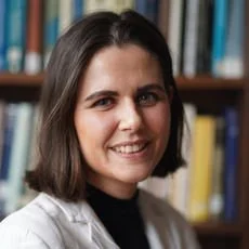 A woman with shoulder-length dark hair smiling in front of a bookshelf filled with colorful books.