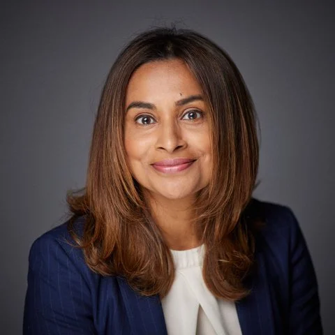 Professional woman with shoulder-length brown hair, wearing a navy blazer and white top, smiling against a gray background.