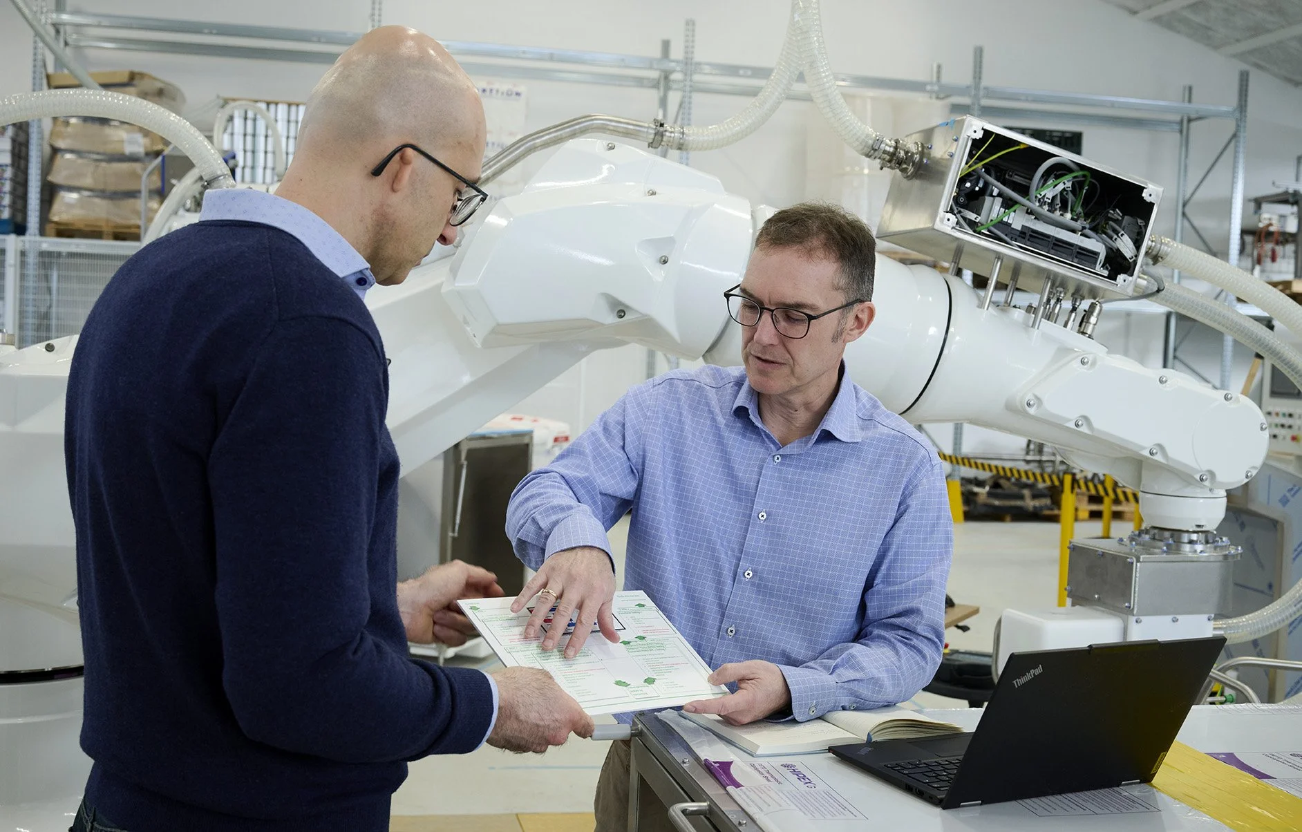 Two men wearing glasses, one with a bald head and the other with short brown hair, discuss documents in an industrial or laboratory setting with mechanical equipment and pipes in the background.