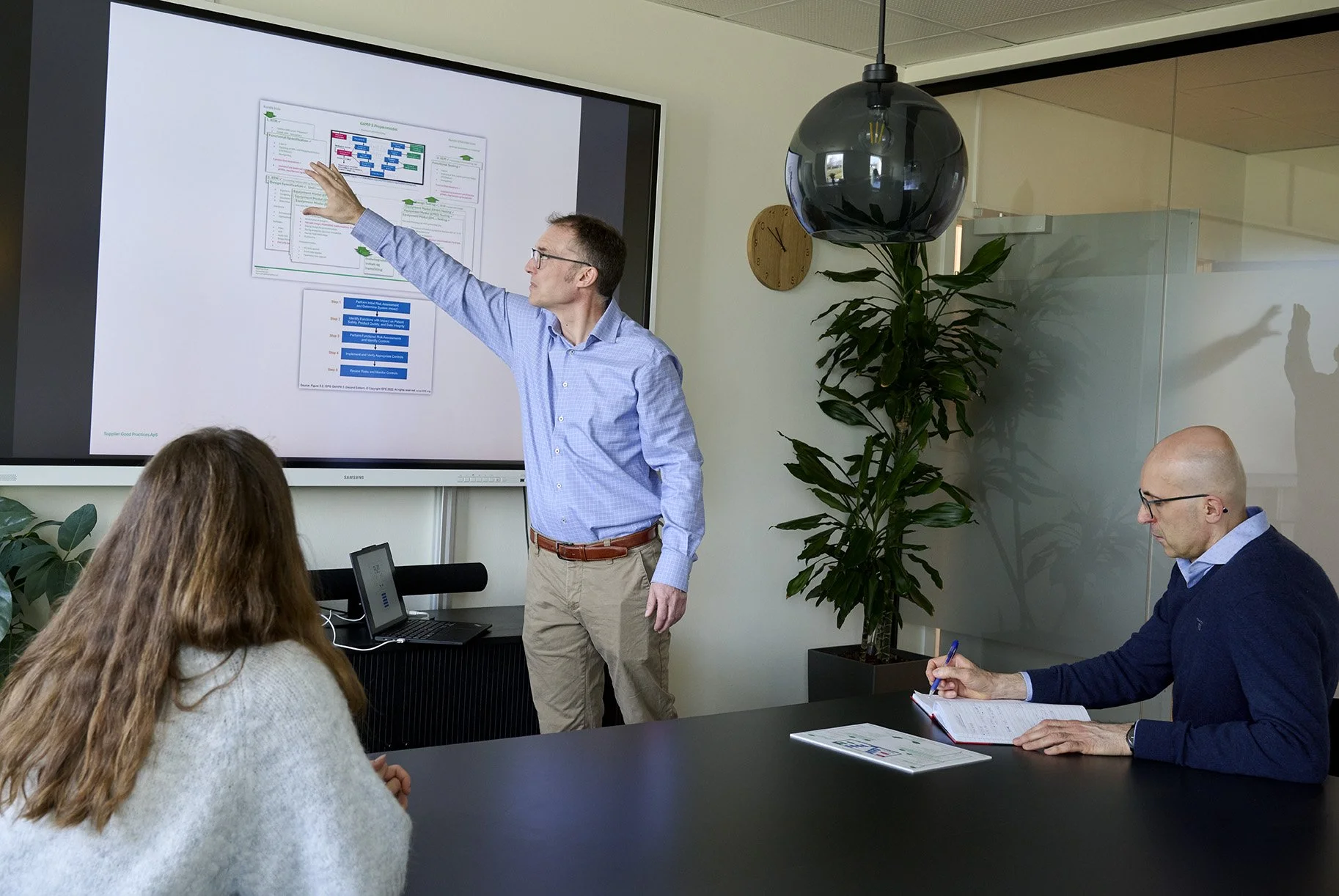 Man giving presentation to group in conference room with large screen, seated participants, laptop, notepad, pen, plants, and wall clock.