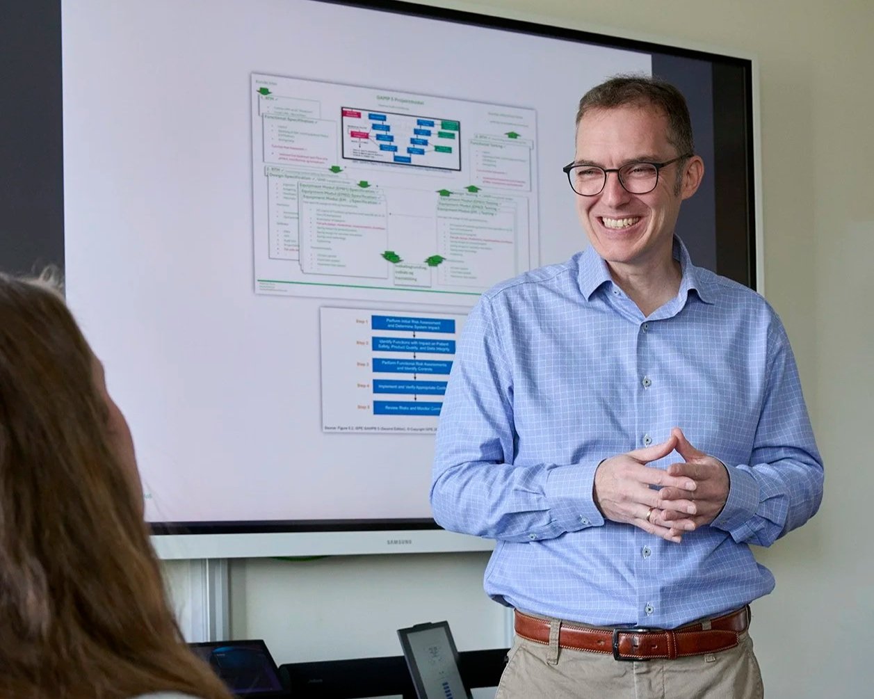 A smiling man wearing glasses and a blue checkered shirt giving a presentation in front of a large screen displaying a flowchart or diagram, with an audience member visible in the foreground.