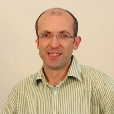 A man with glasses, short brown hair, and a slight smile, wearing a light green striped shirt, against a plain beige background.