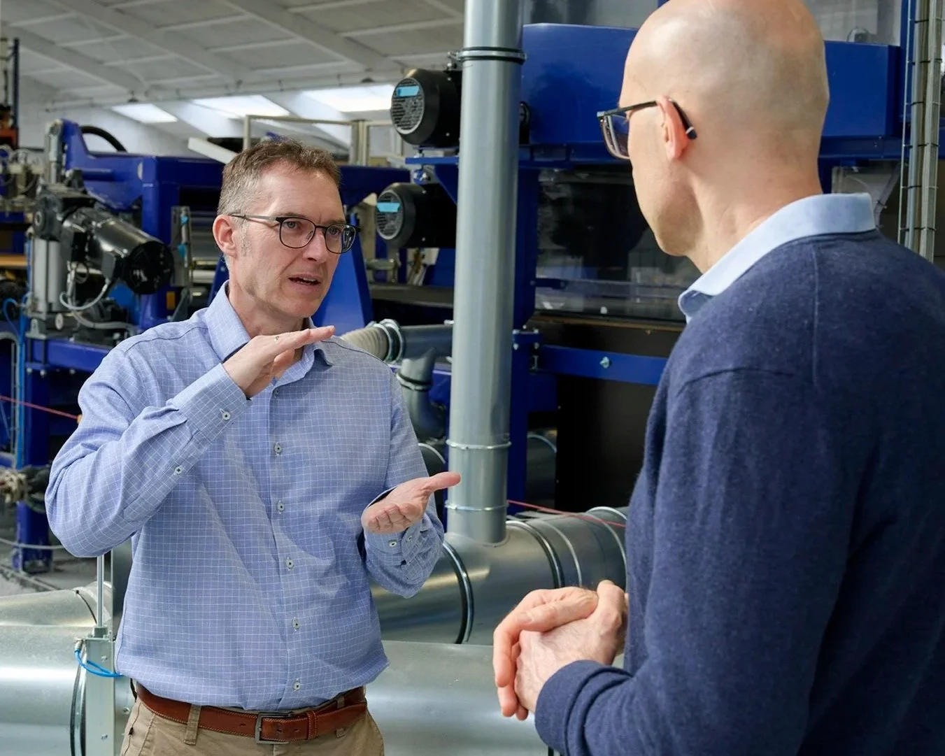 Two men in conversation in an industrial setting, with blue machinery and pipes in the background. One man is gesturing with his hands, while the other listens.