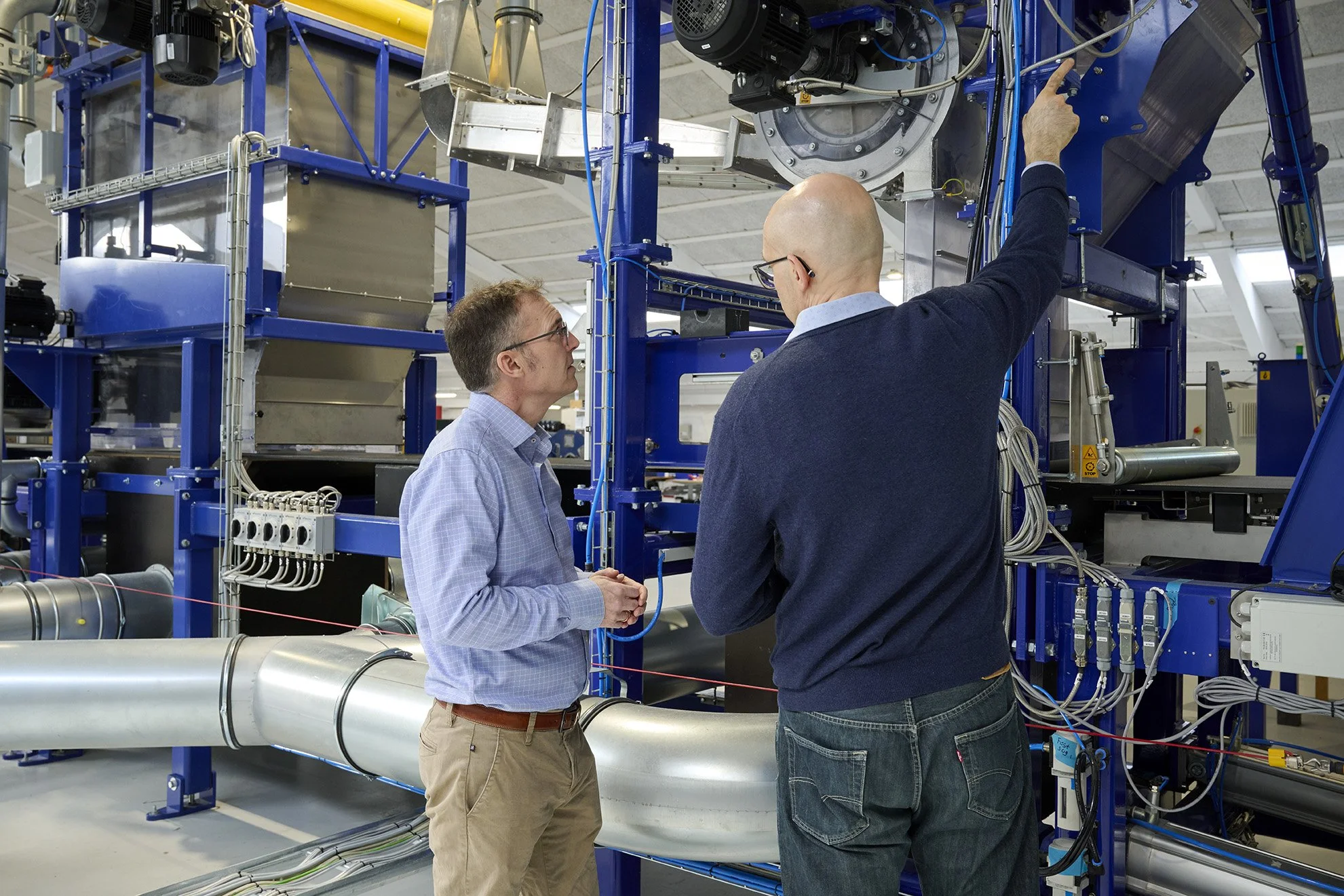 Two men are speaking in front of a large industrial machine with blue framing and various pipes, wires, and mechanical components in a factory or research facility.