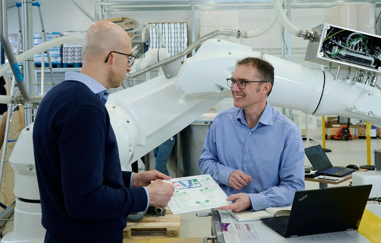 Two men in a laboratory or industrial setting, one holding a paper with charts, engaged in conversation with a large robotic arm and equipment in the background.